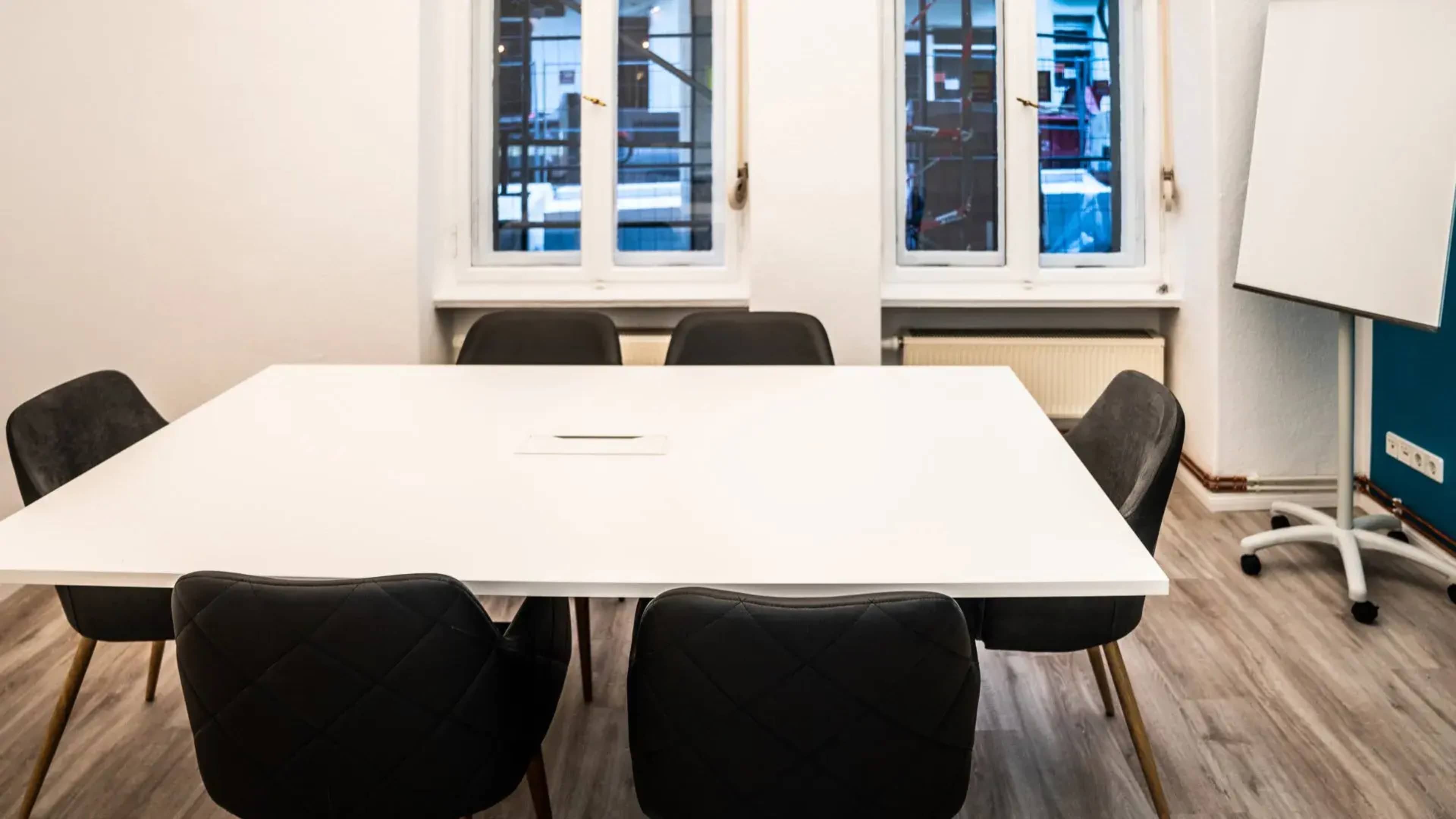 A large white conference table surrounded by six black chairs is positioned in a room with two windows showing an outdoor view.