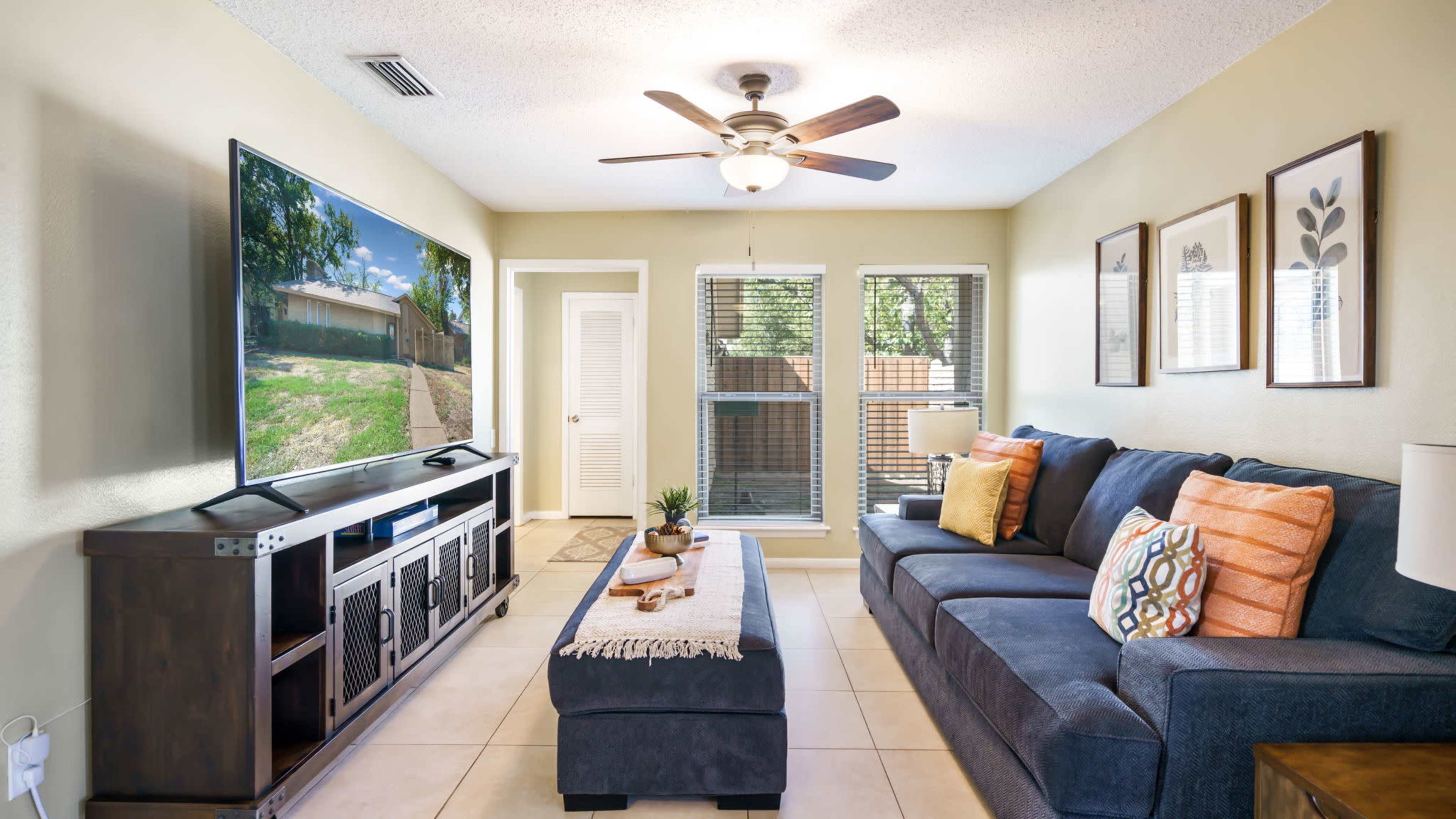 The living room features a sectional sofa, a large television mounted on a stand, and windows allowing natural light, with a coffee table and decorative items arranged centrally.