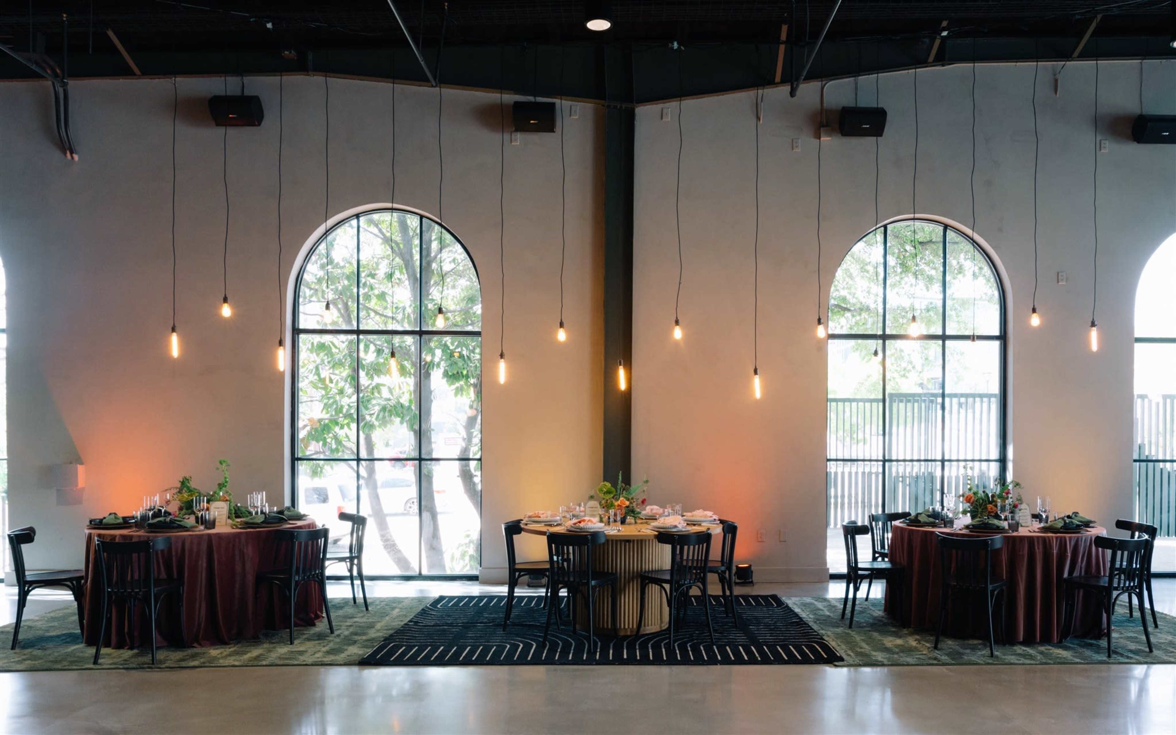 The image shows a dining area with two round tables elegantly set, surrounded by large arched windows and illuminated by hanging pendant lights.