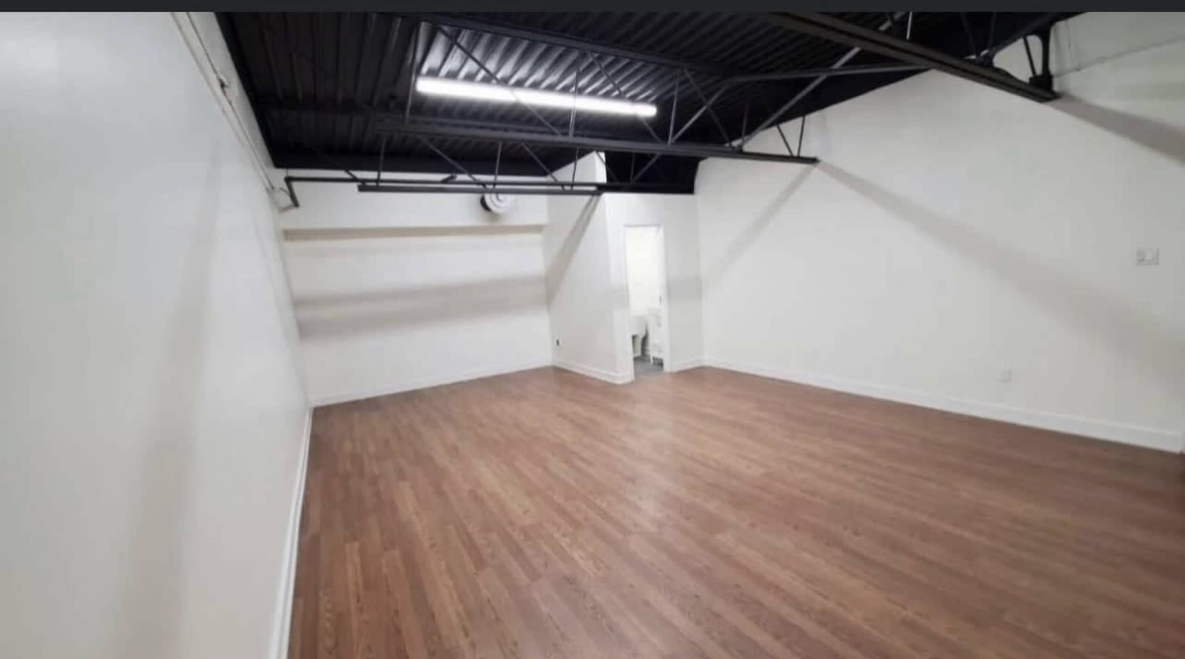 A large, empty room with hardwood flooring and white walls, featuring exposed black beams in the ceiling.