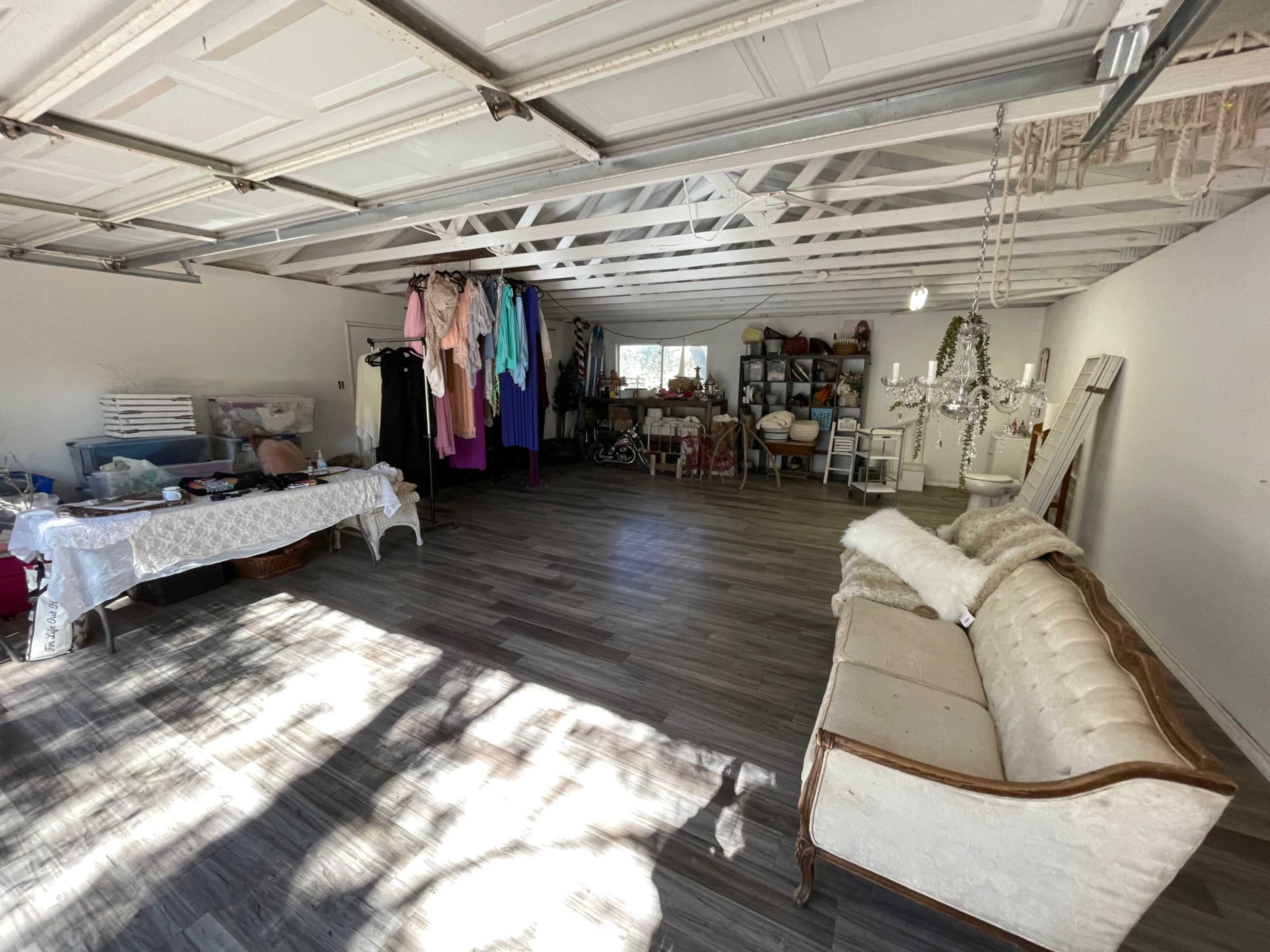 A spacious garage interior with a variety of clothing hanging on racks, a table covered with items, and a vintage couch positioned along one wall.