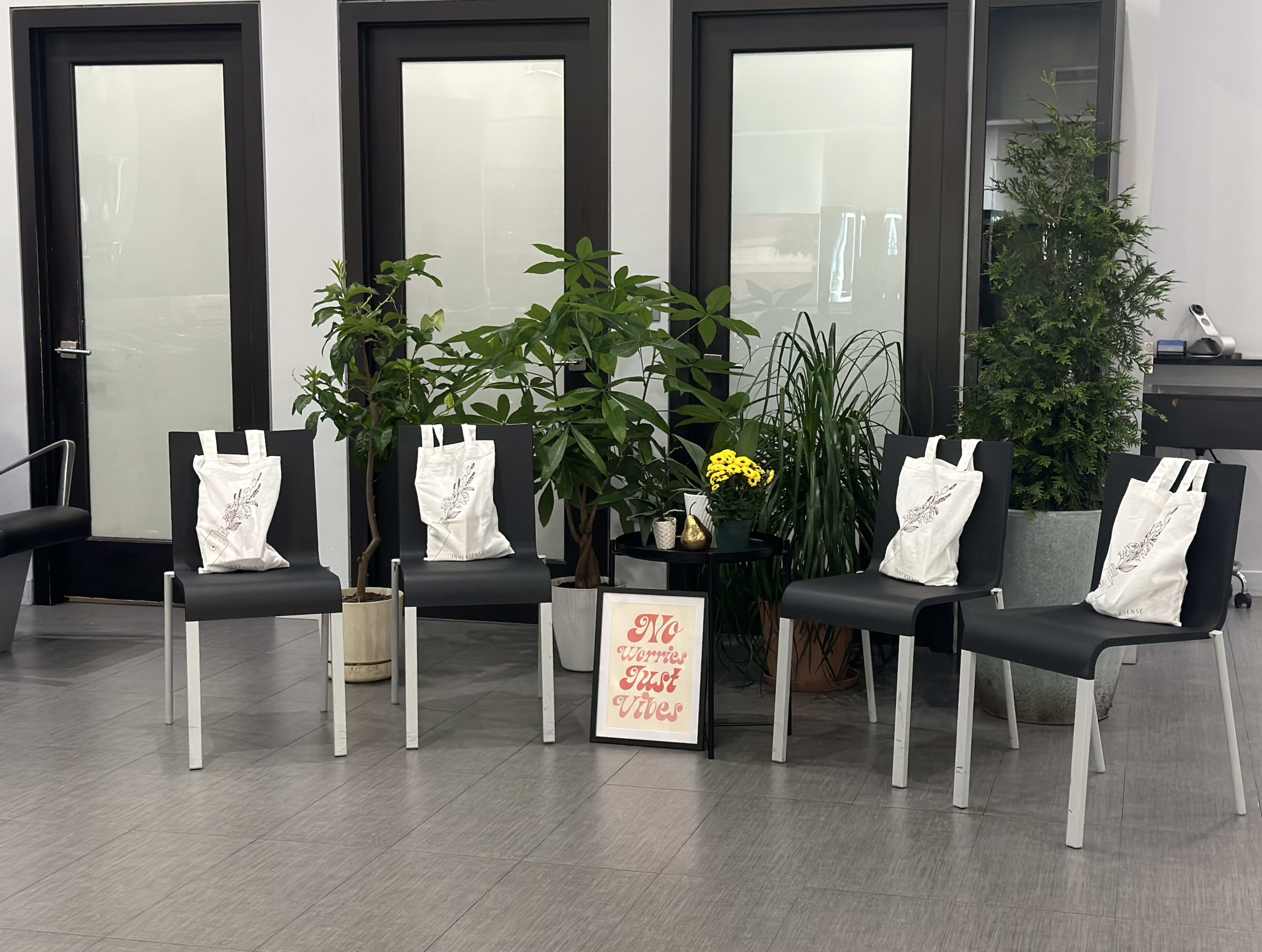 Four empty black chairs with tote bags resting on them, surrounded by potted plants and a sign that reads "No Turmoil Just Vibes."