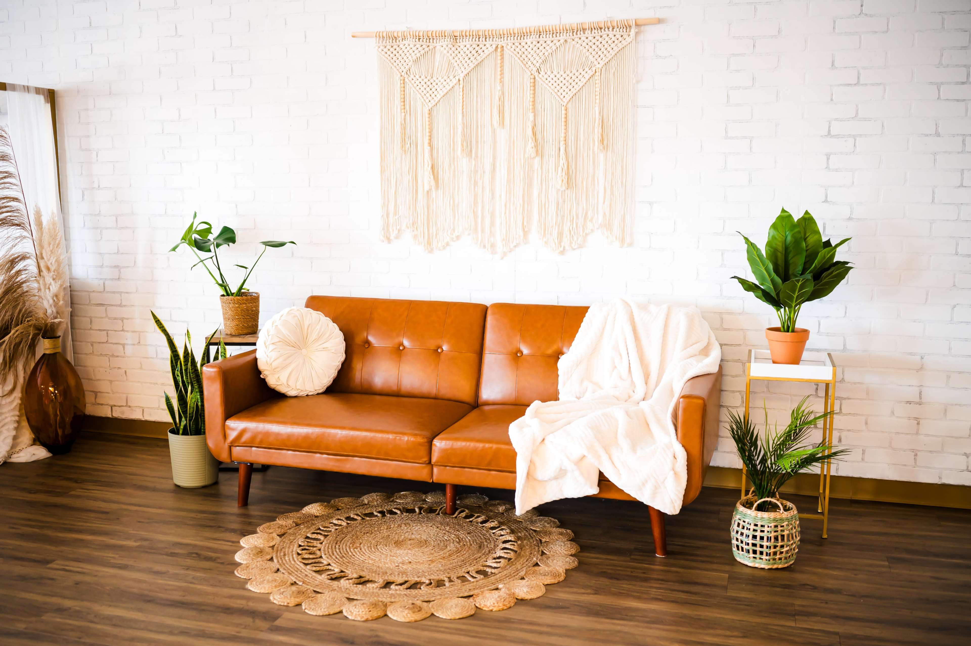 A leather sofa is placed against a white brick wall, surrounded by various plants and a woven area rug.