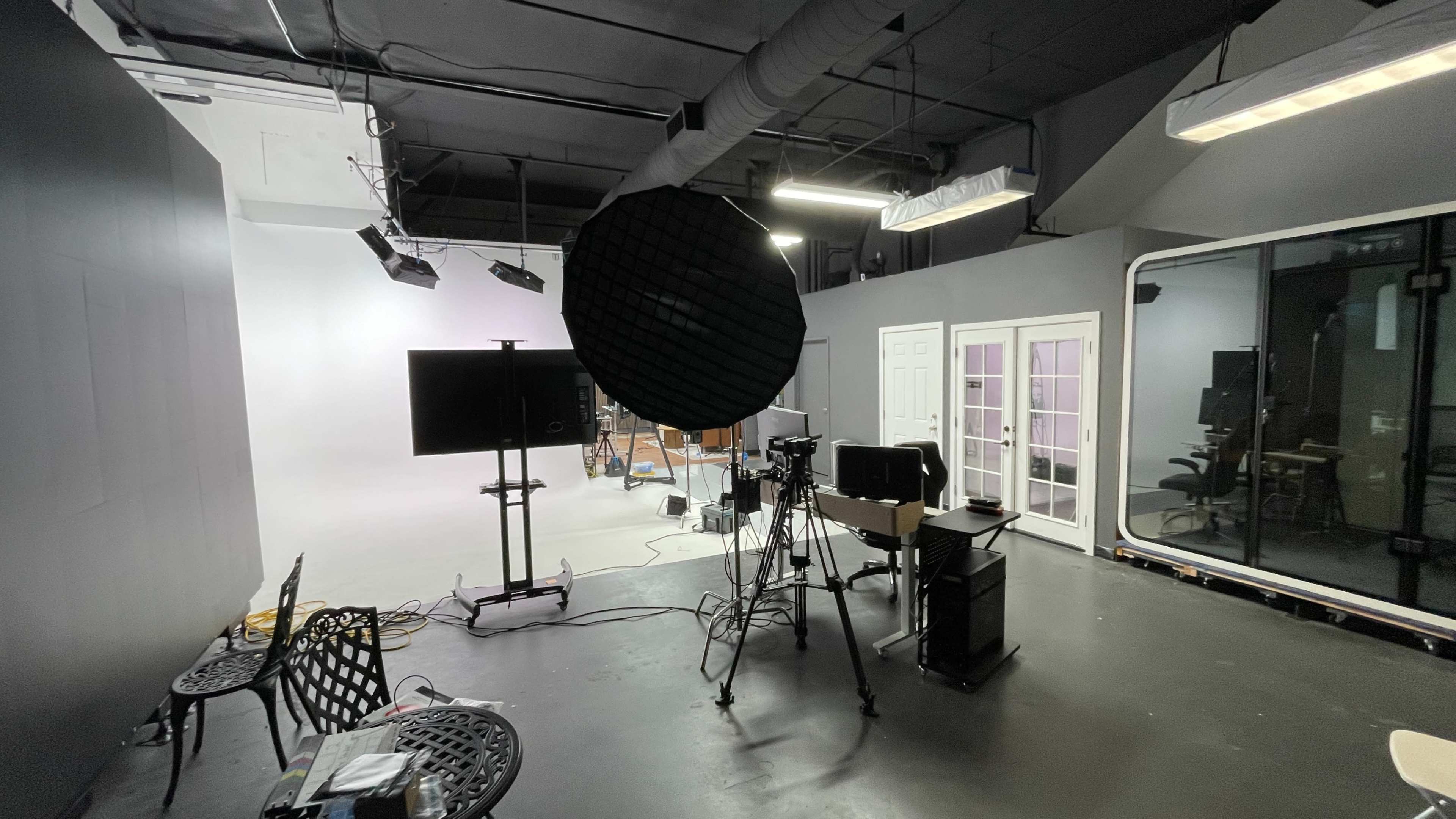 The image shows a photography or video production studio with various equipment, including lighting, cameras, and a monitor, set against gray walls.