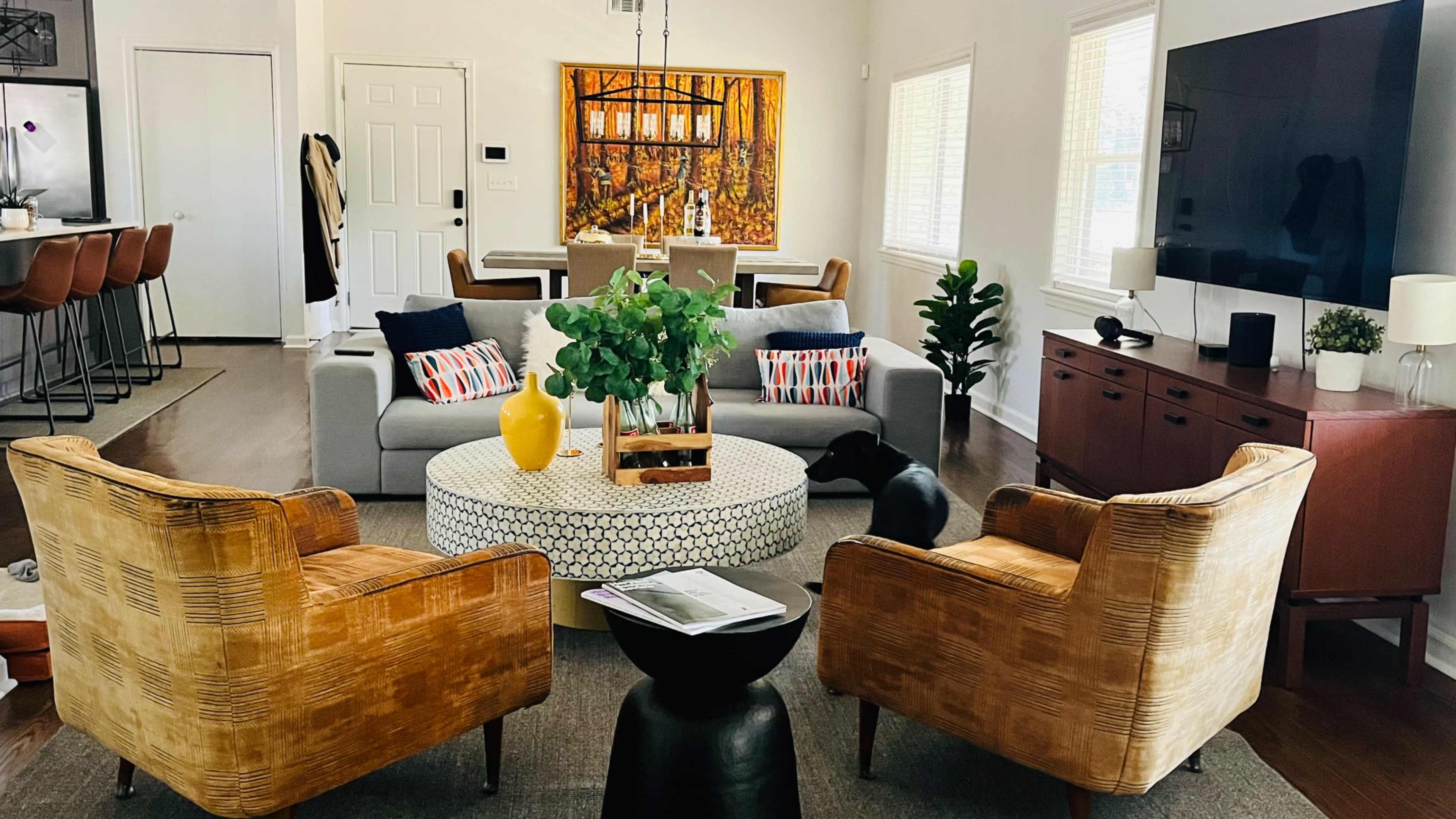 The image shows a modern living room with a gray sofa, two wicker armchairs, a round coffee table with a vase, and a large television on one wall.
