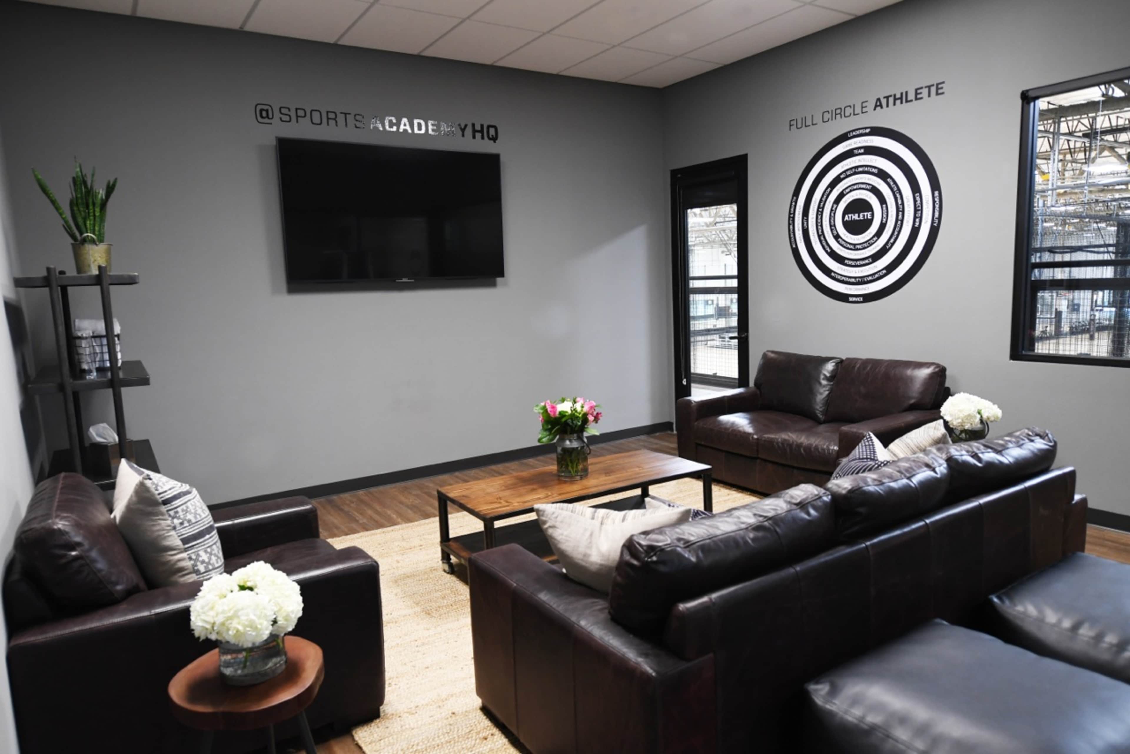 A modern lounge area with two dark leather sofas, a coffee table, a television mounted on the wall, and decorative wall art, all situated in a bright room with large windows.