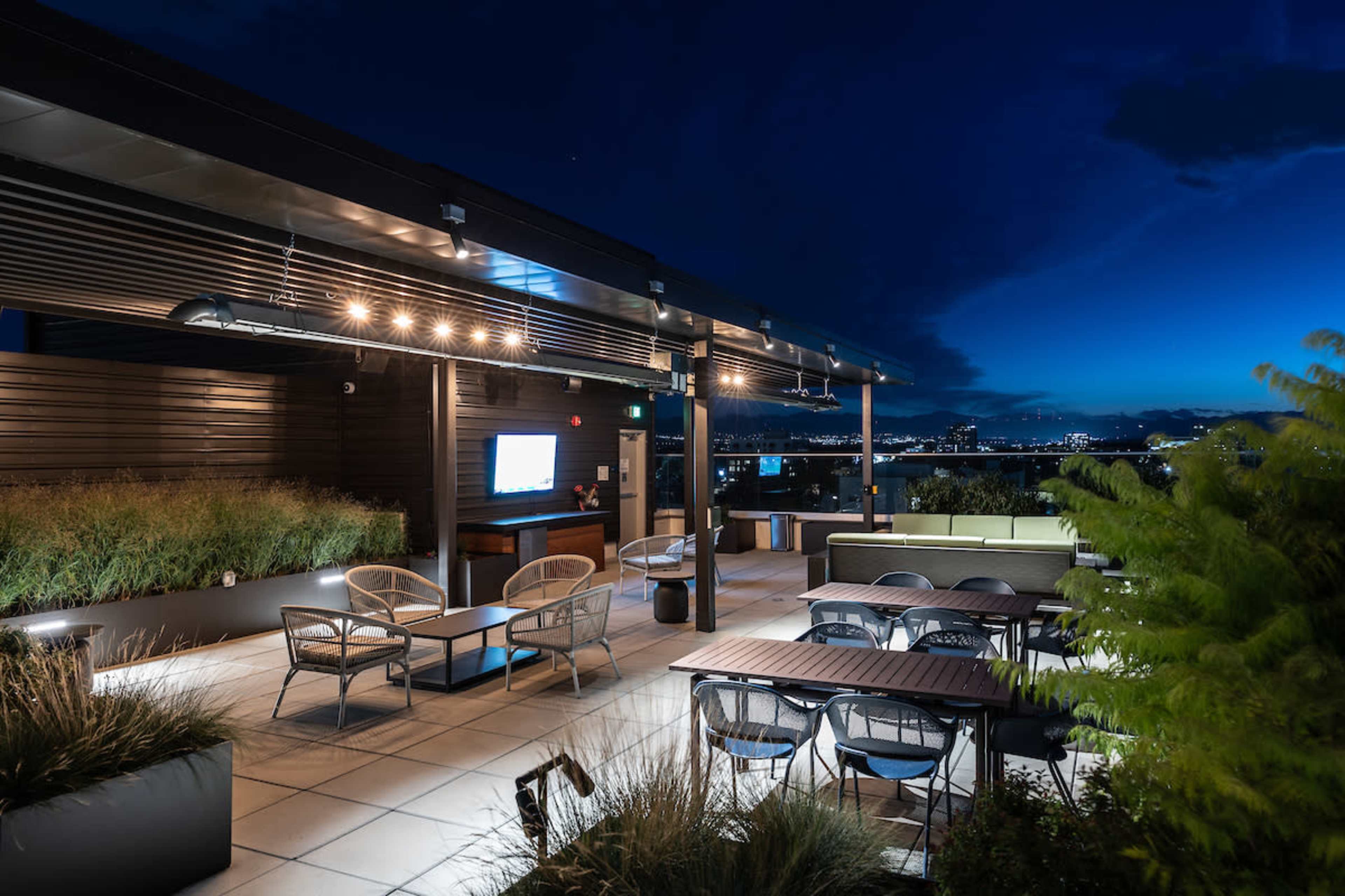 A rooftop terrace features seating areas, planters with tall grass, and ambient lighting against a twilight sky.
