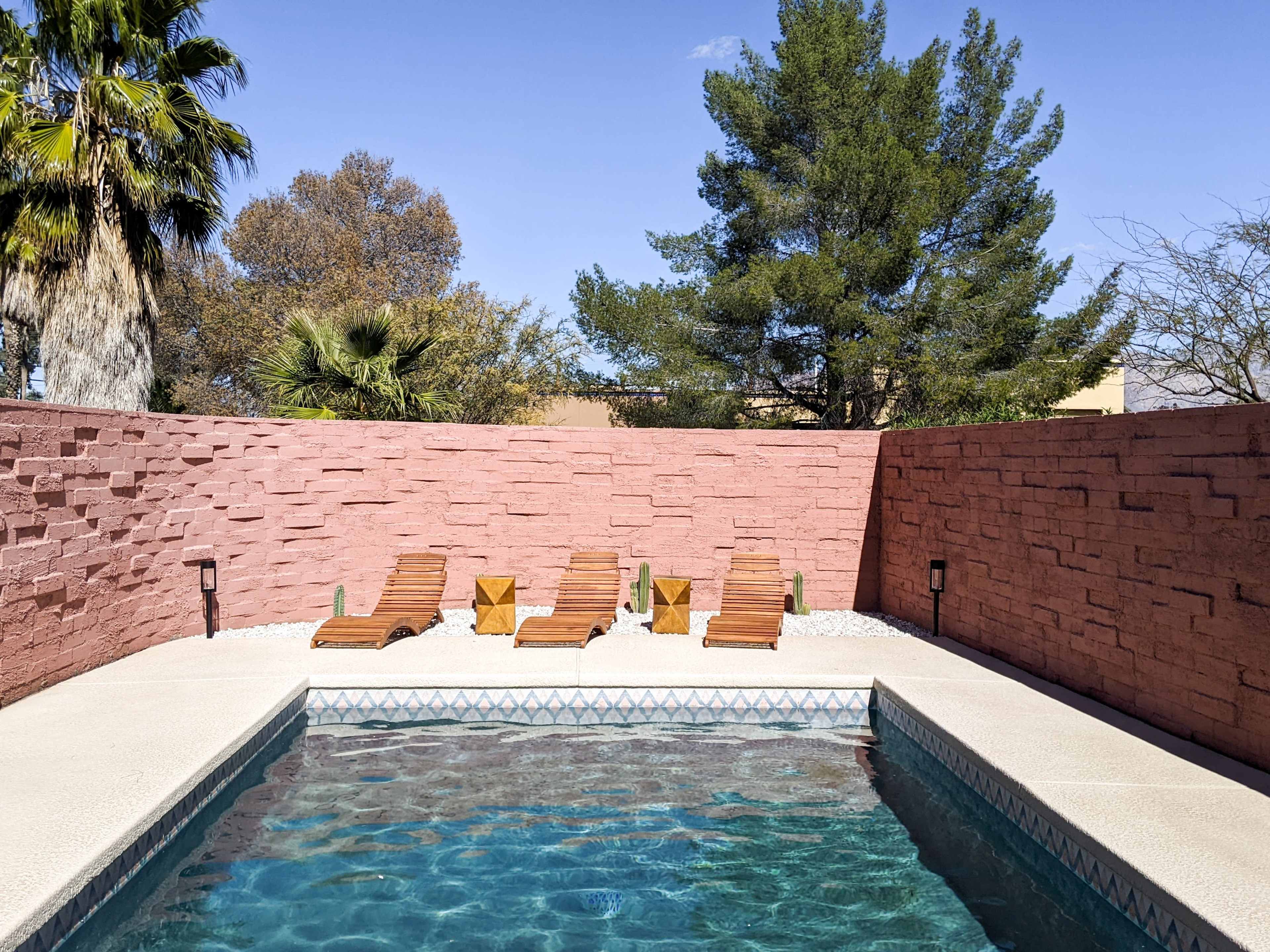 A swimming pool surrounded by a pink stone wall and four wooden lounge chairs beside it.
