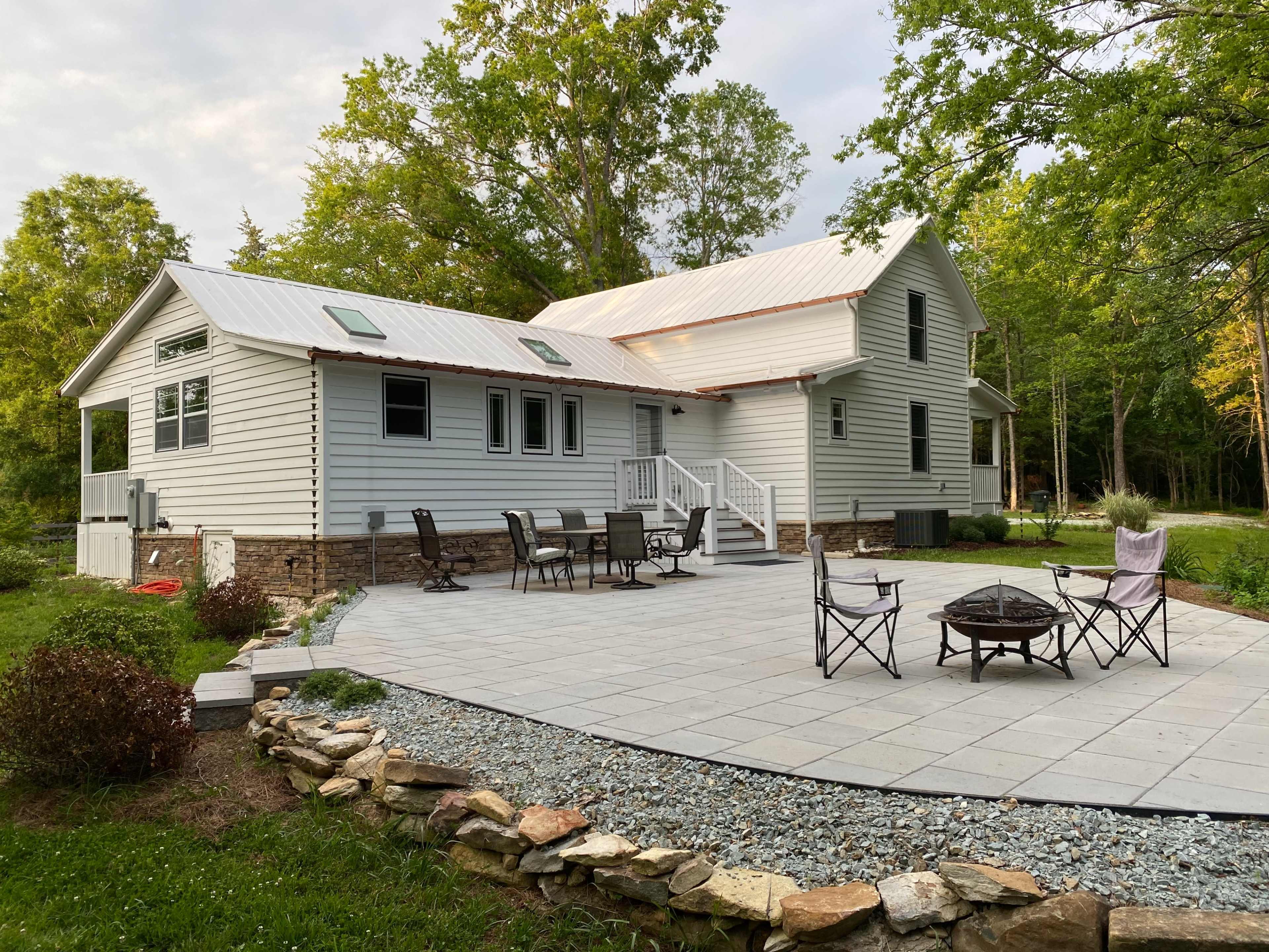 A two-story house with a metal roof features a stone patio and outdoor seating area surrounded by trees.