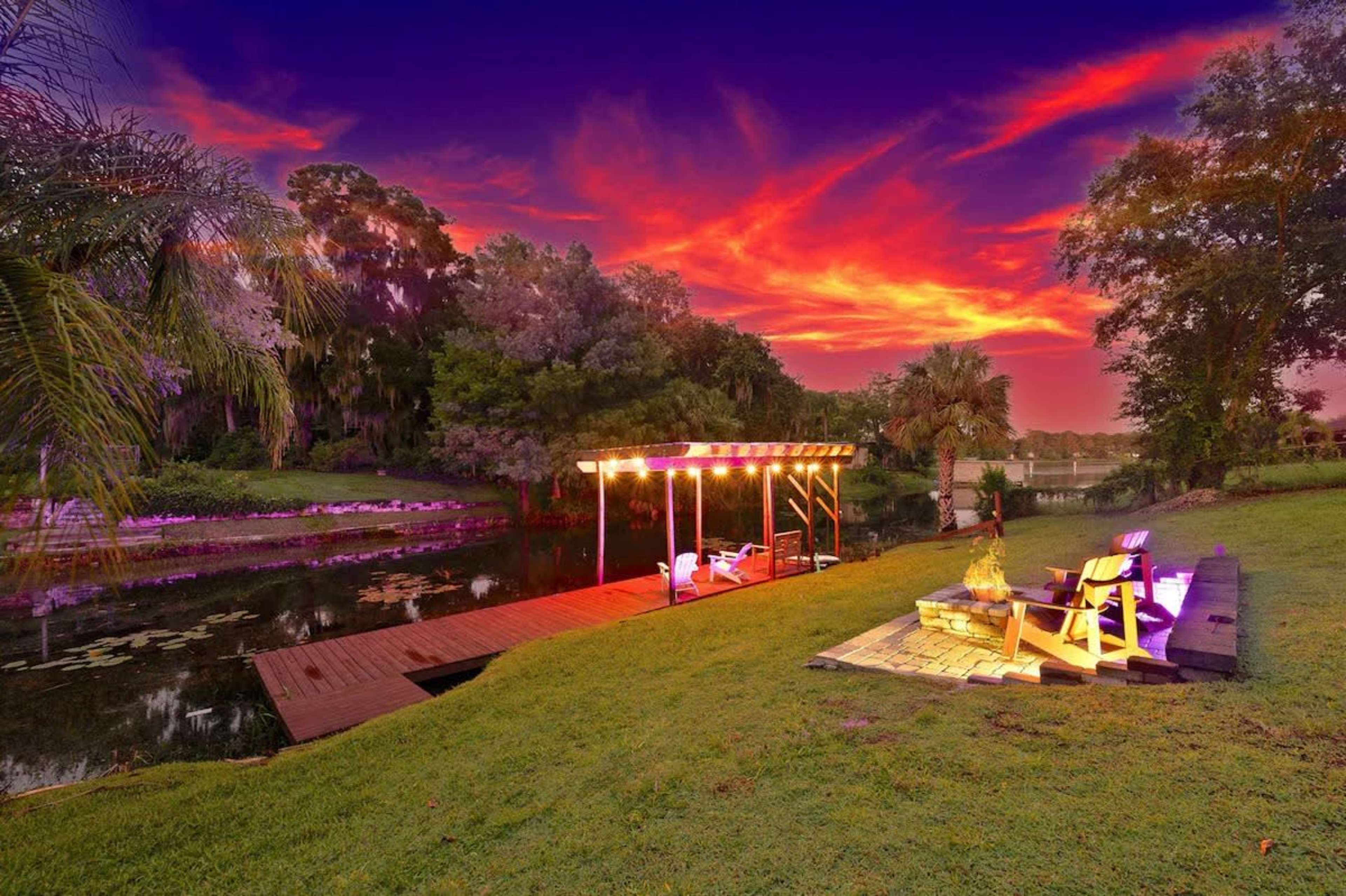 A wooden dock with lounge chairs overlooks a body of water under a vibrant sunset sky filled with clouds.