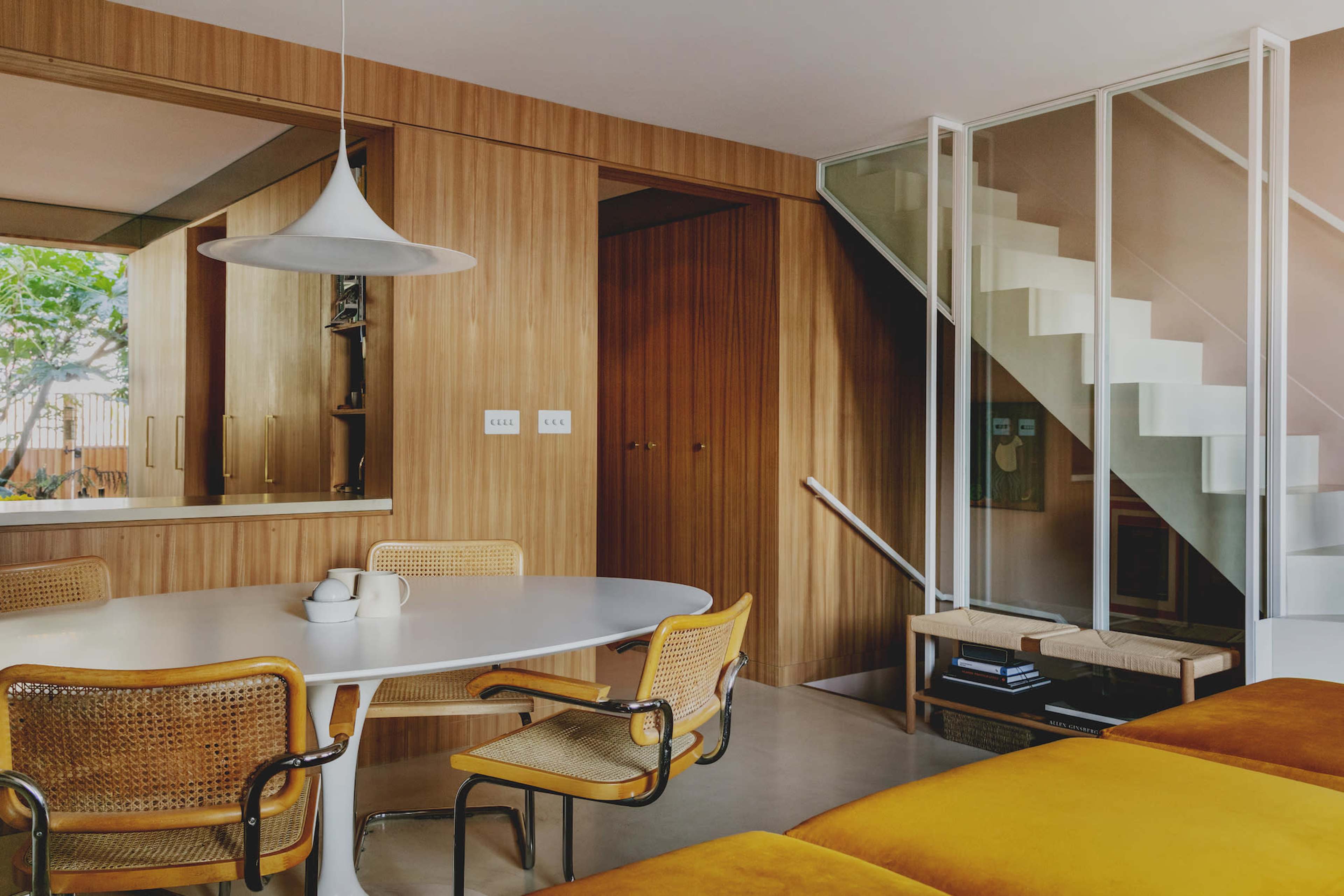 A modern interior featuring a dining area with a round table and wicker chairs, adjacent to a staircase with a glass enclosure, all set against wooden paneling.