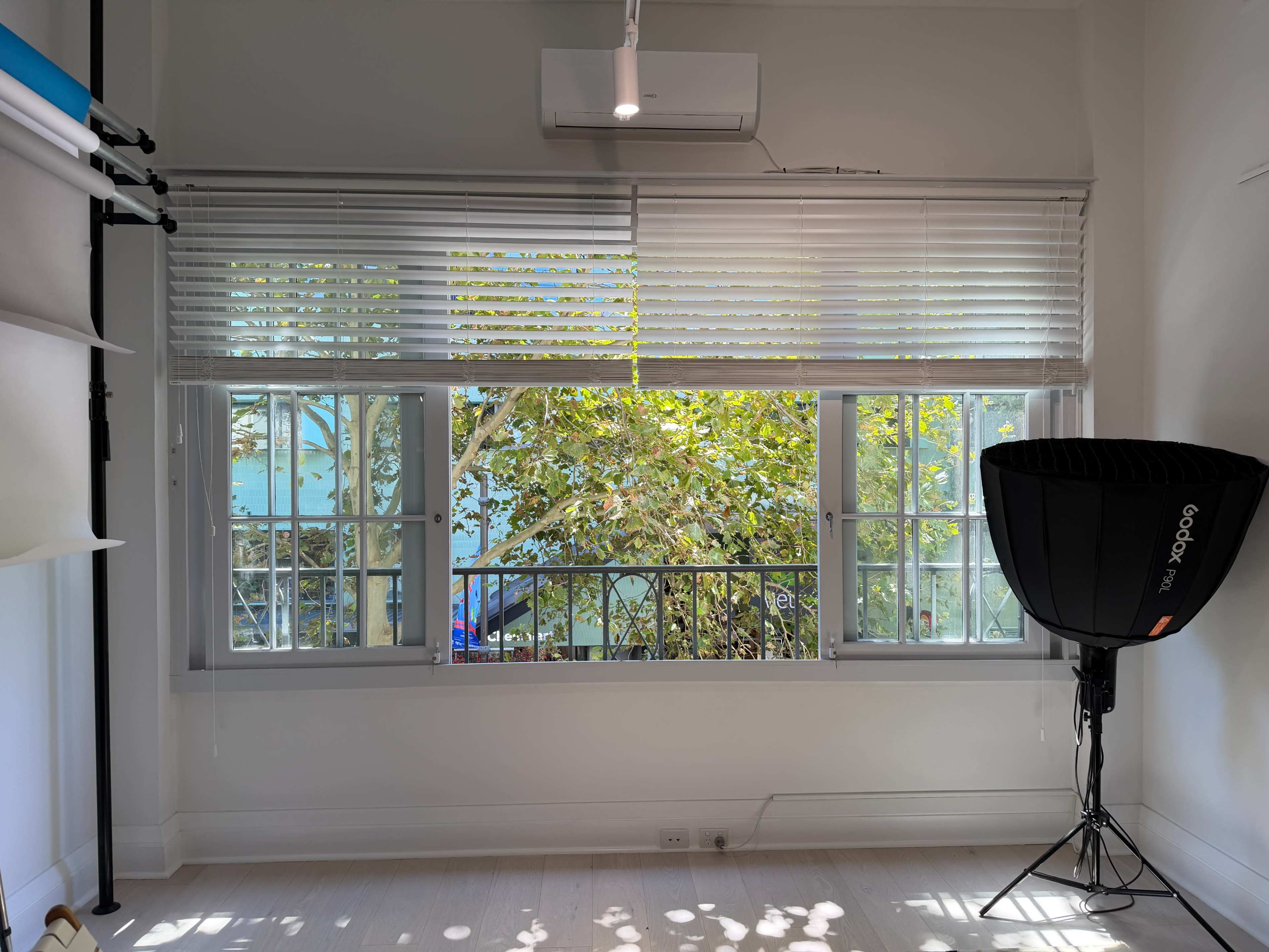 The image shows a bright room with large windows overlooking trees and a body of water, accompanied by a photography light stand in the corner.