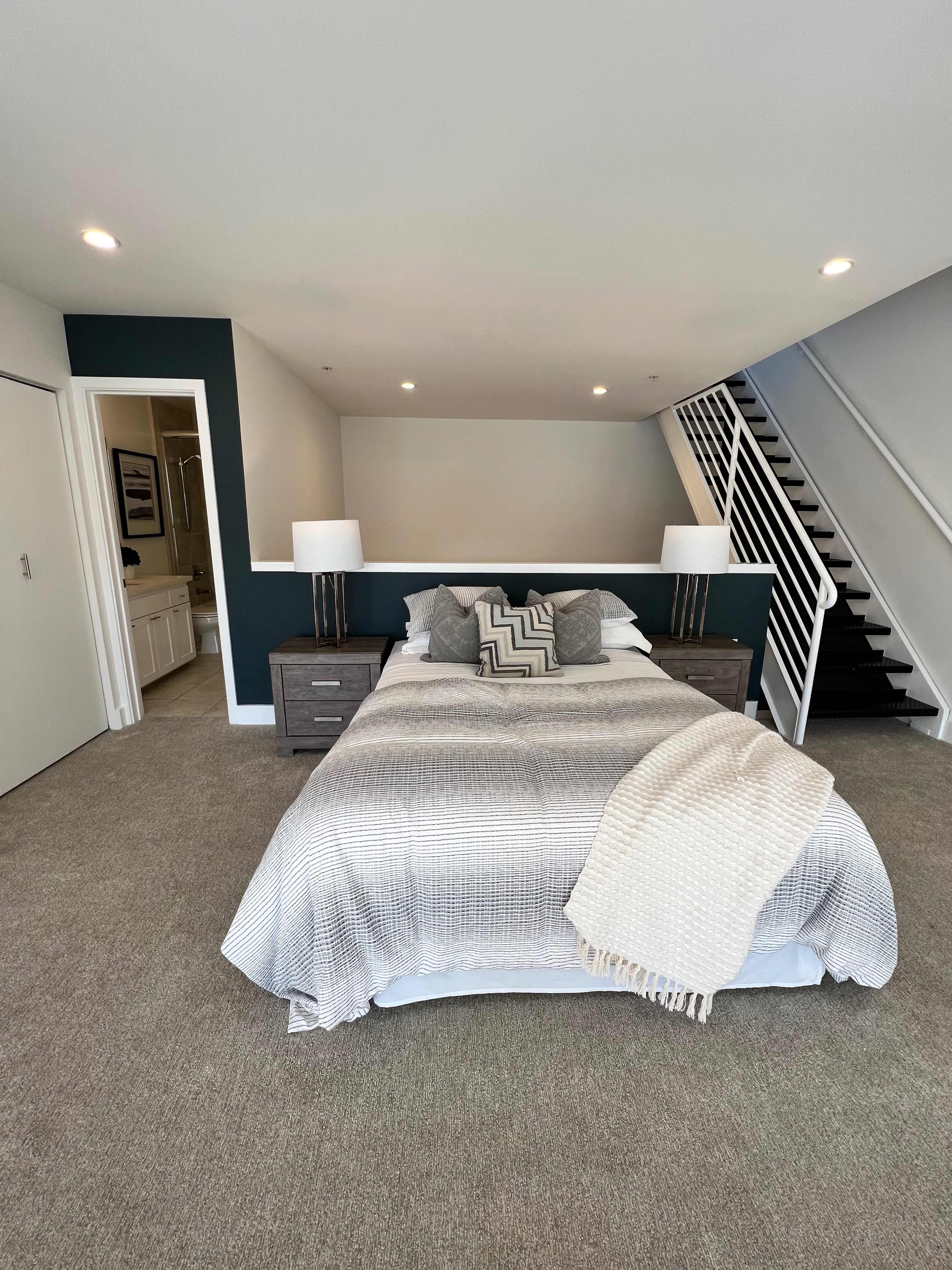 A modern bedroom with a double bed, two bedside lamps, and a staircase visible in the background.