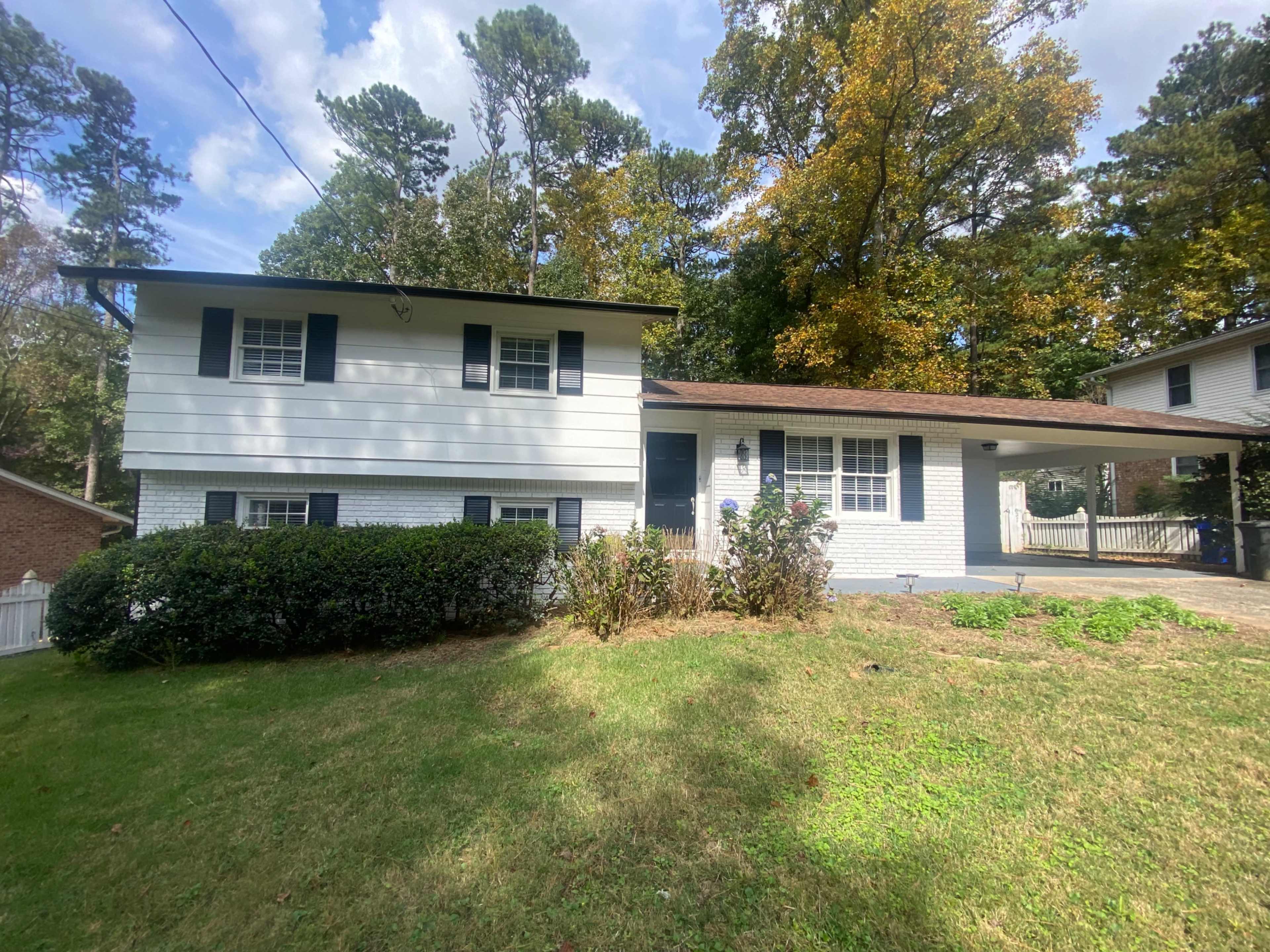 A two-story white house with a brown roof and black shutters is surrounded by green grass and trees.