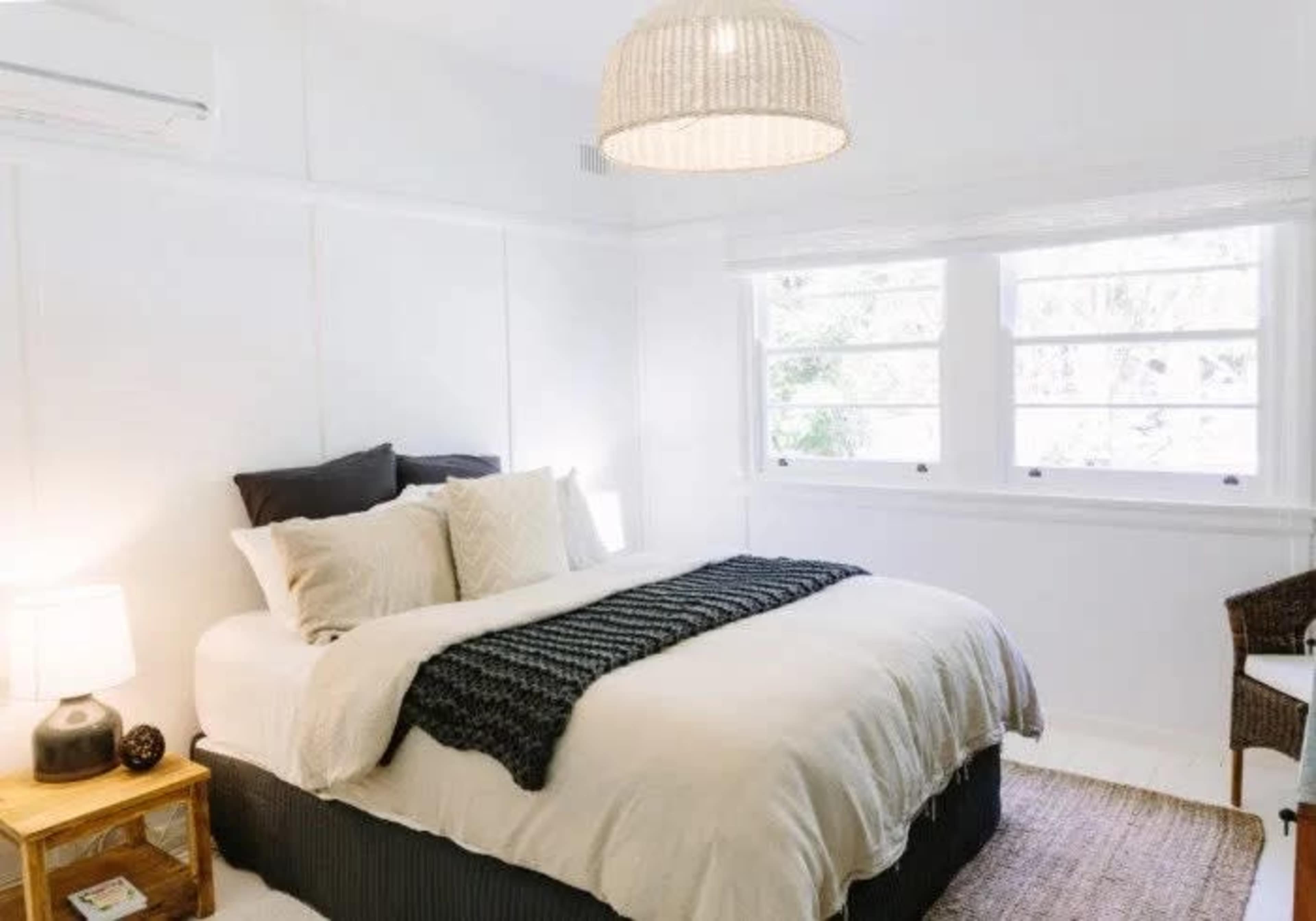 A cozy bedroom features a neatly made bed with cream-colored bedding and a woven throw, accompanied by a bedside lamp and wicker chair against a white-paneled wall.