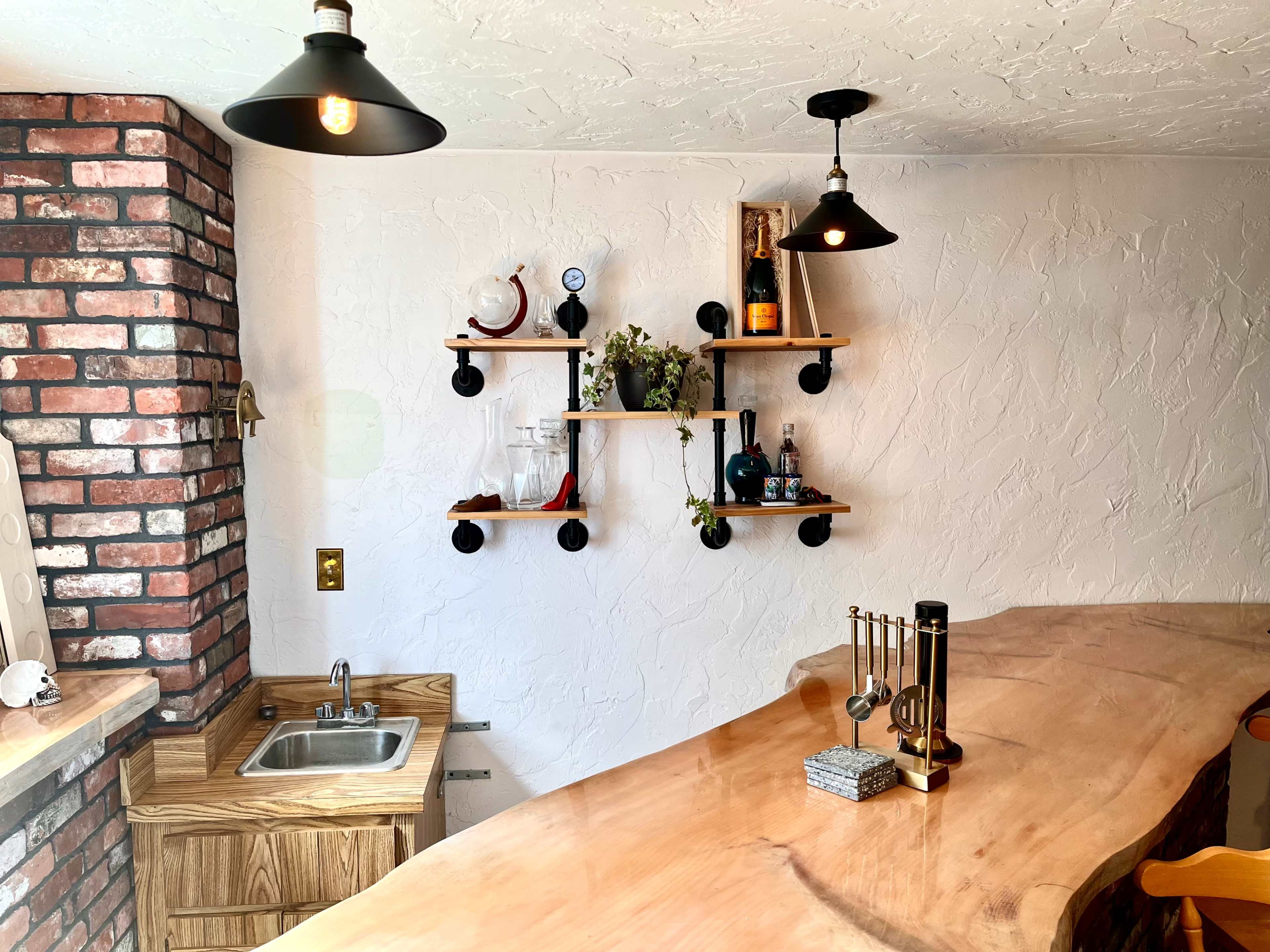 The image shows a kitchen area featuring a wooden bar counter, a small sink, and shelves displaying bottles and decorative items against a textured white wall and exposed brick.