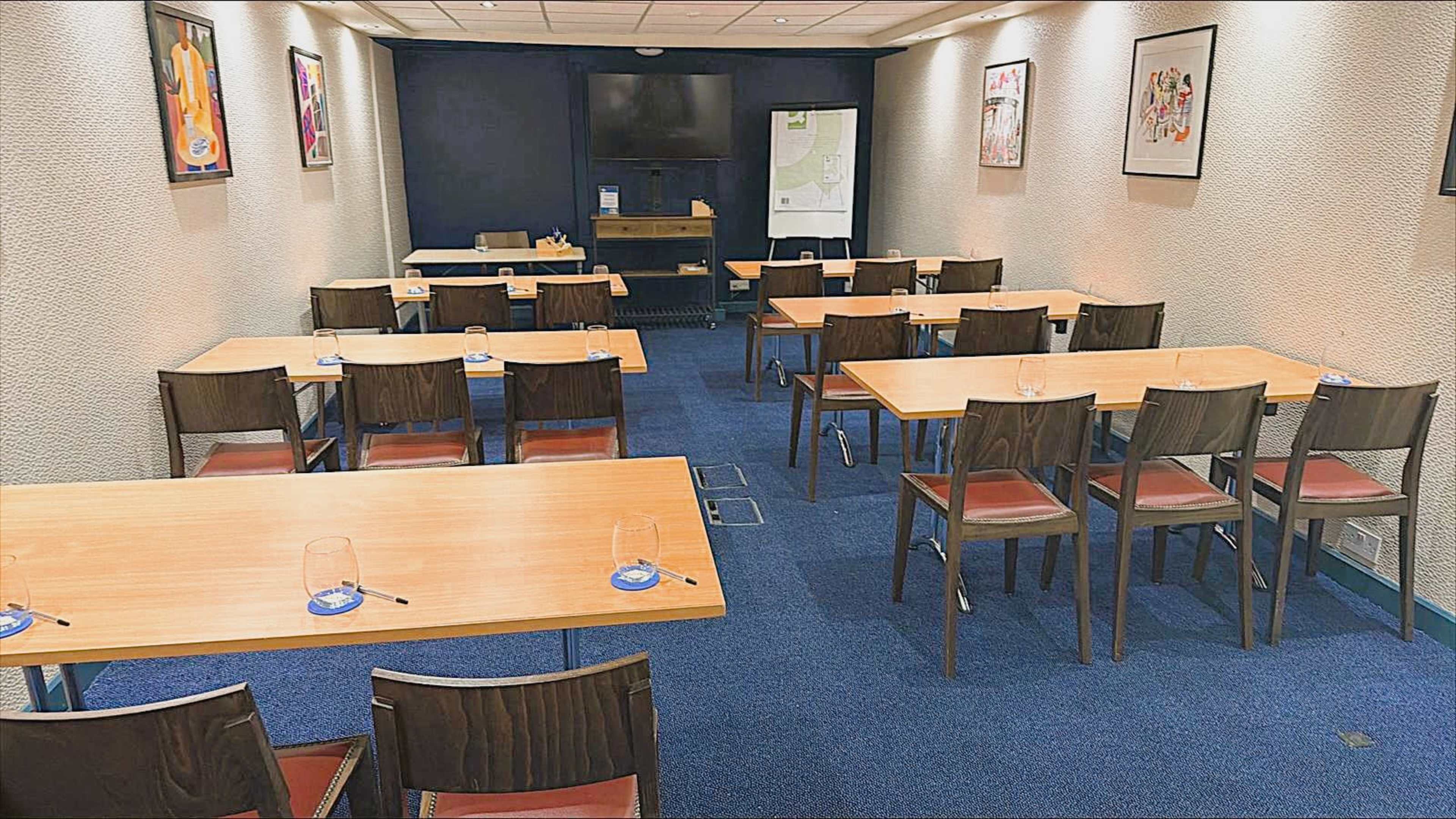 The image shows a conference room with wooden tables and chairs arranged in a grid format, with a television on the wall and artwork displayed.