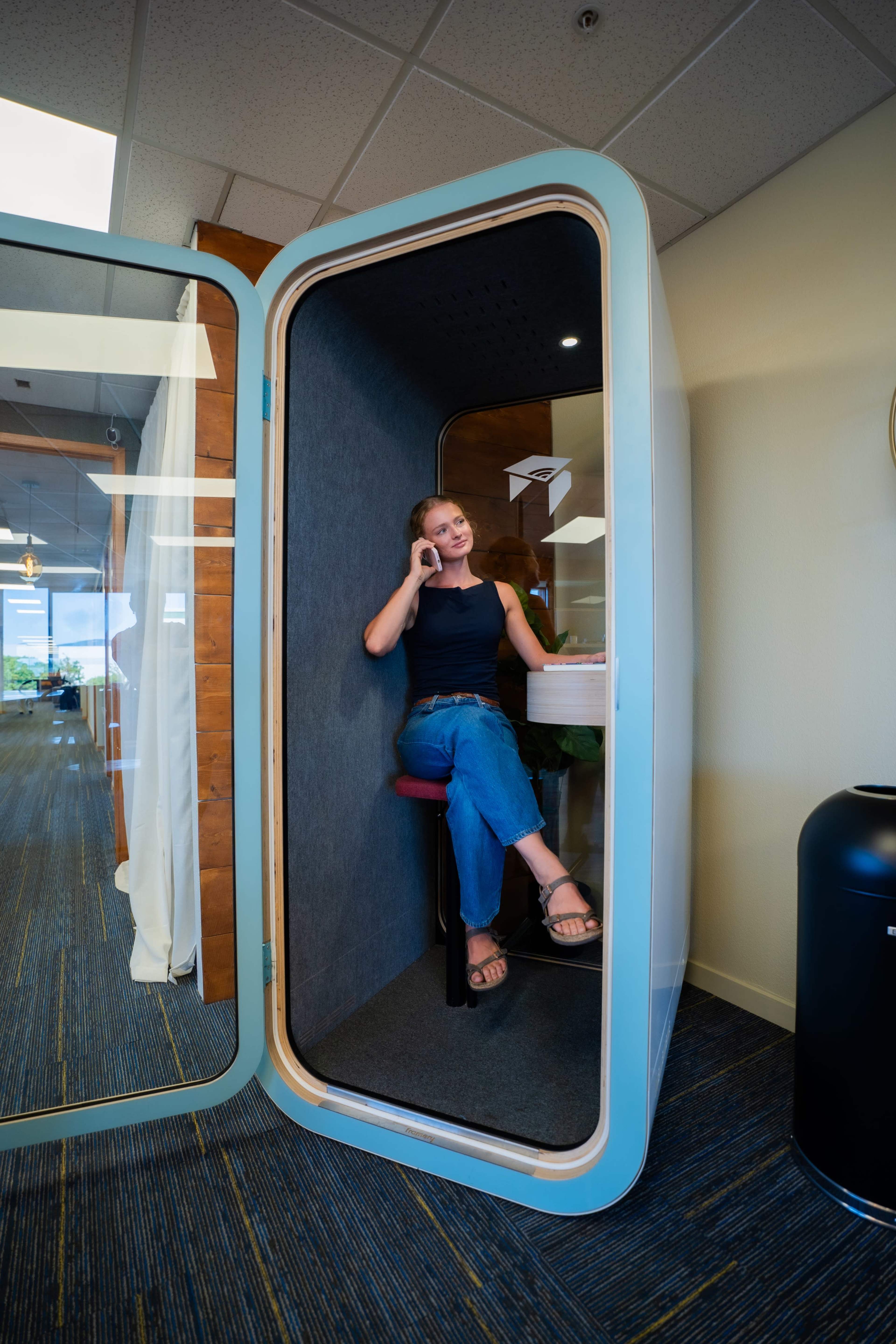 A woman sits inside a soundproof booth with a phone to her ear, surrounded by modern office decor.