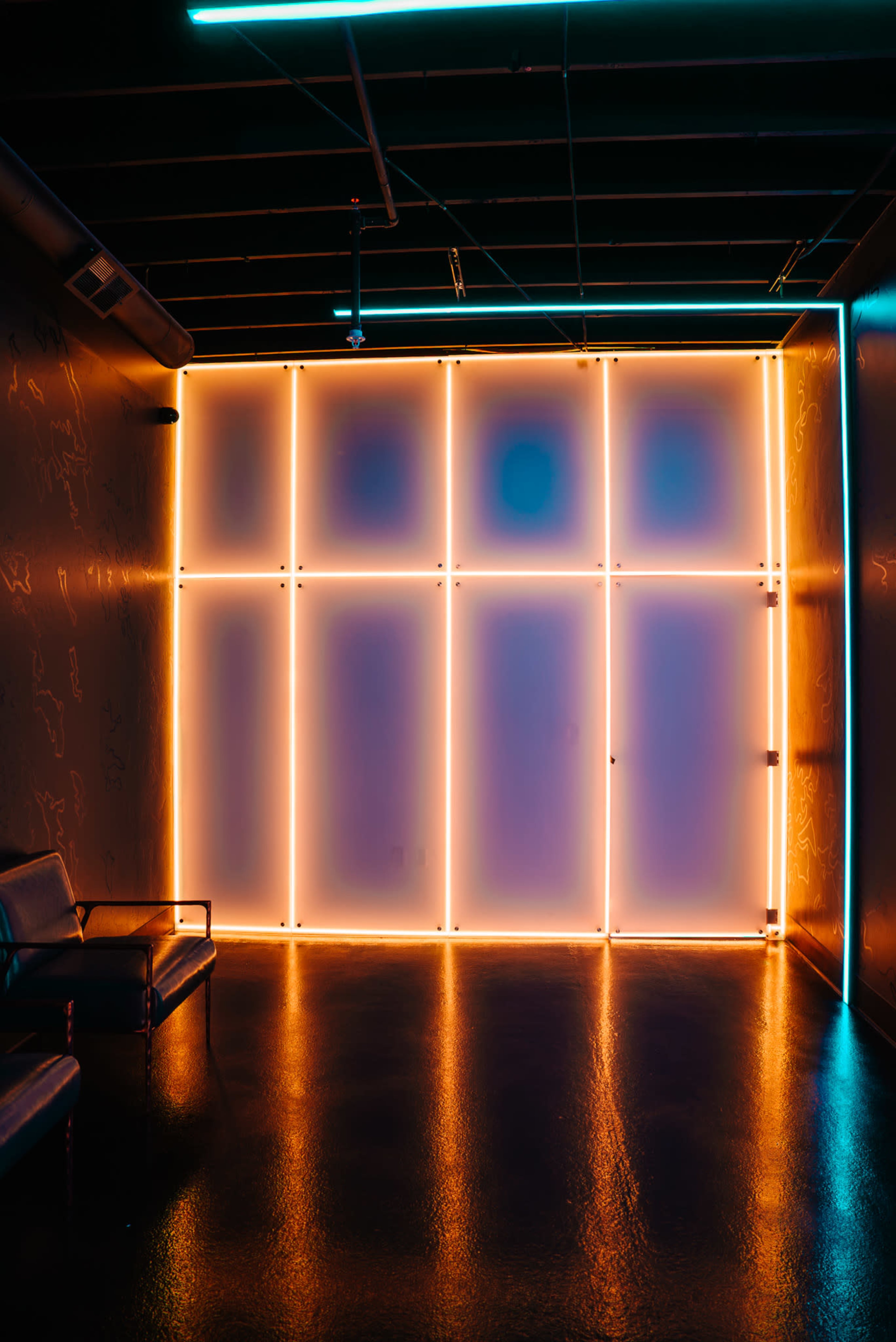 A dimly lit room features a wall illuminated by vibrant neon lights, creating a striking contrast with the dark floor and minimalist furniture.