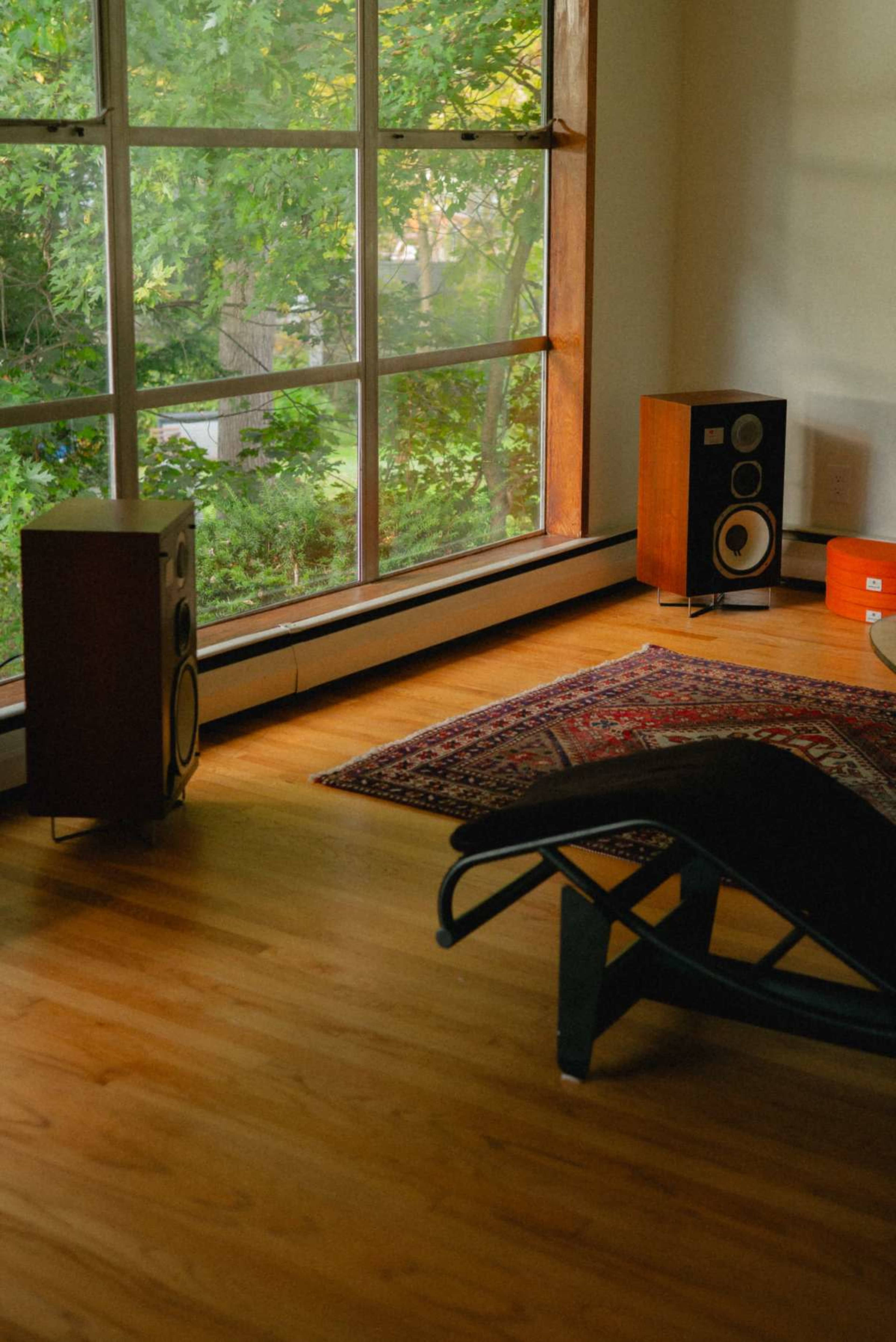 The image shows a cozy room with large windows, a wooden floor, two speakers, and a patterned rug.