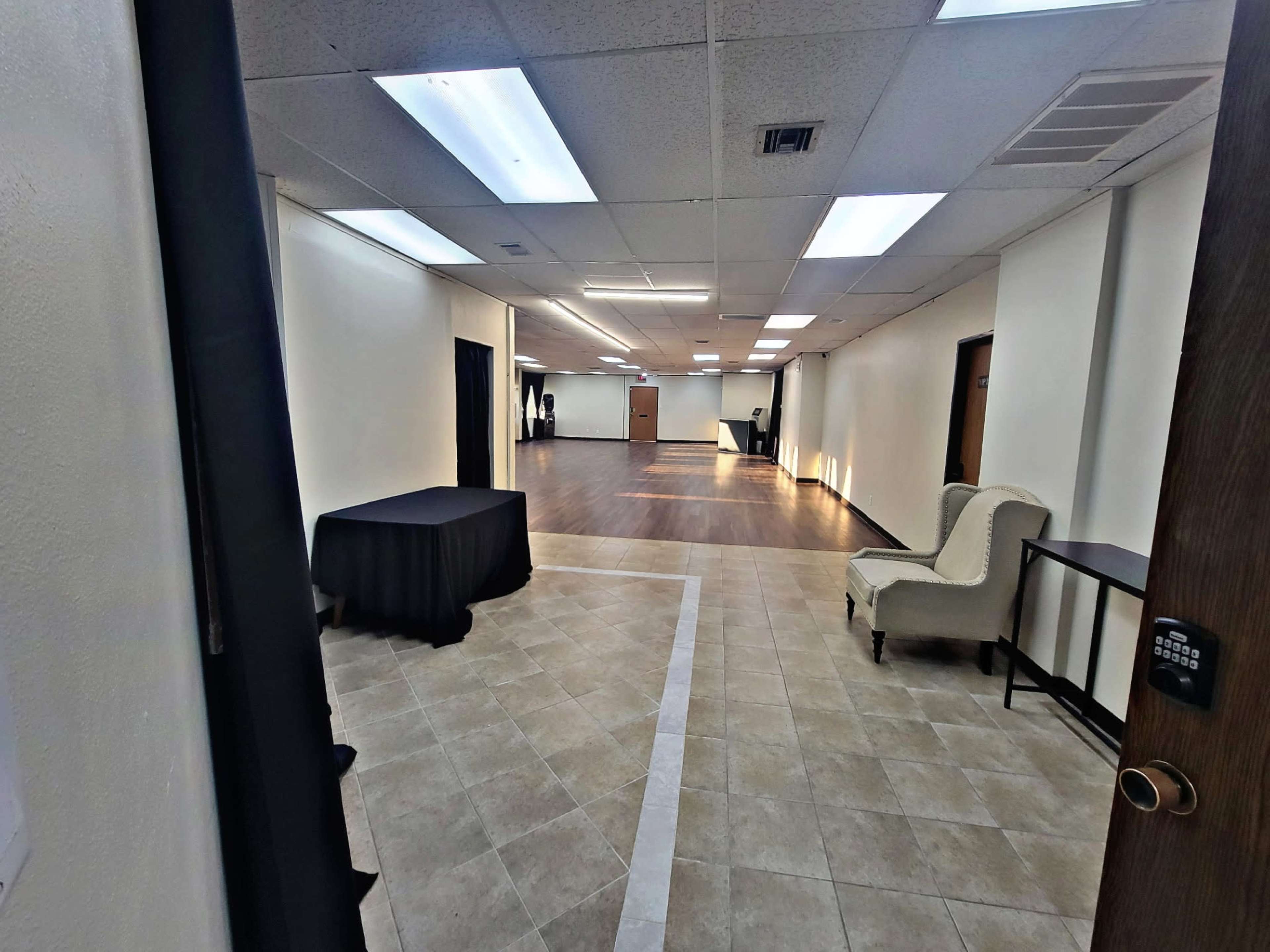 The image shows a spacious, well-lit interior room with tiled flooring, featuring a large table, a chair, and multiple doorways.