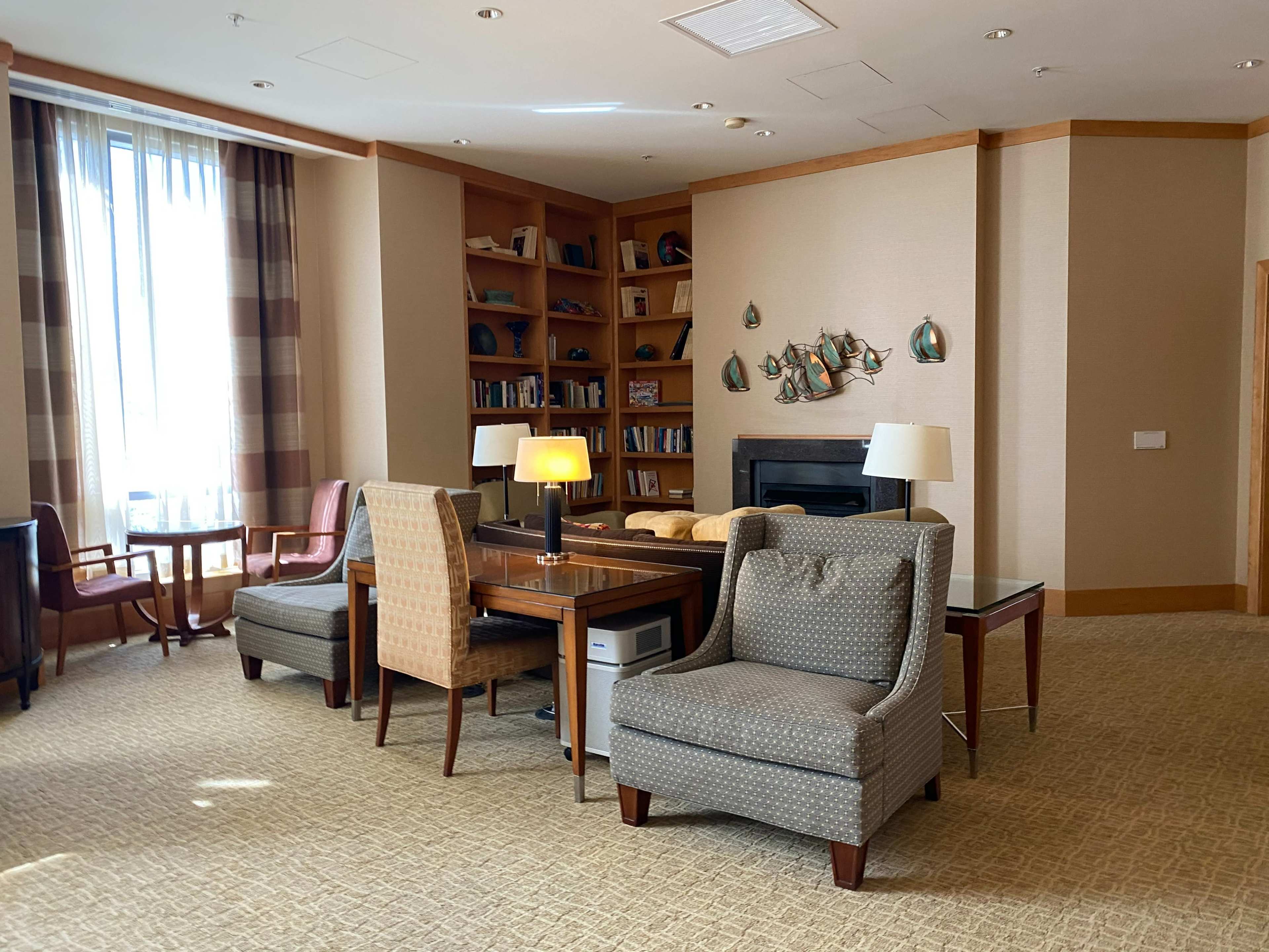 A well-lit lounge area with a bookshelf, comfortable chairs, and a table near a fireplace.