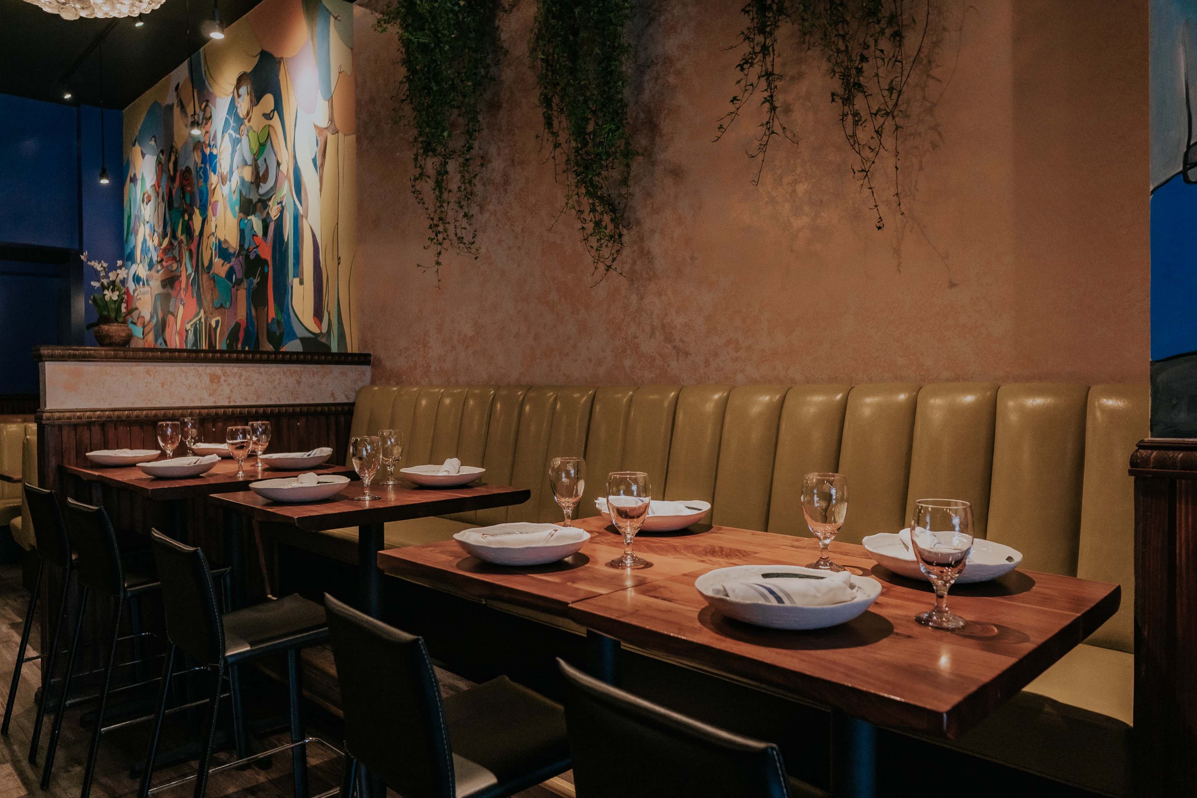 The image shows a dining area with wooden tables set with plates and glasses, surrounded by yellow upholstered benches and a colorful mural on the wall.