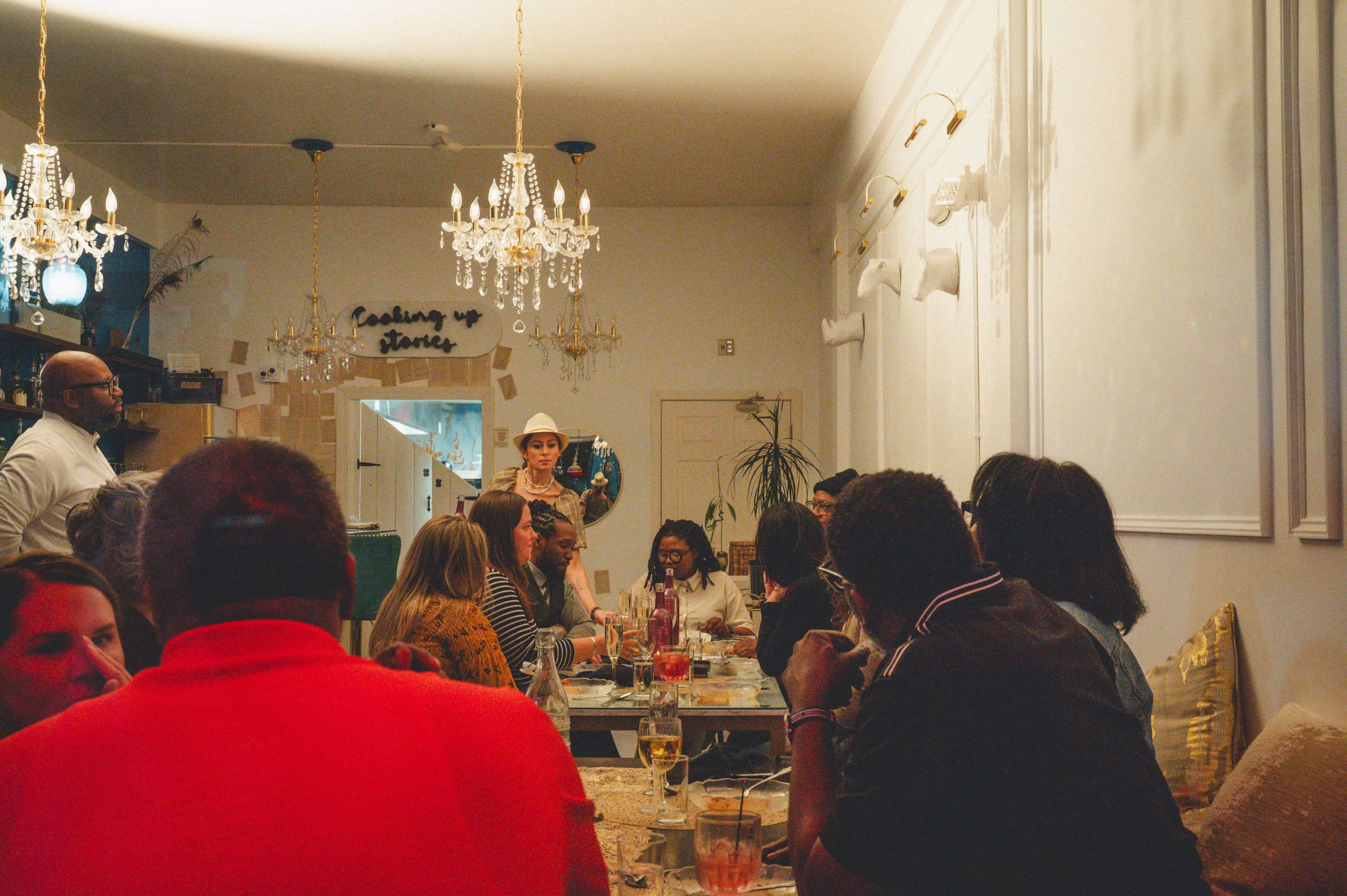 A woman stands in front of a table of diners in a restaurant filled with decorative chandeliers and wall art.