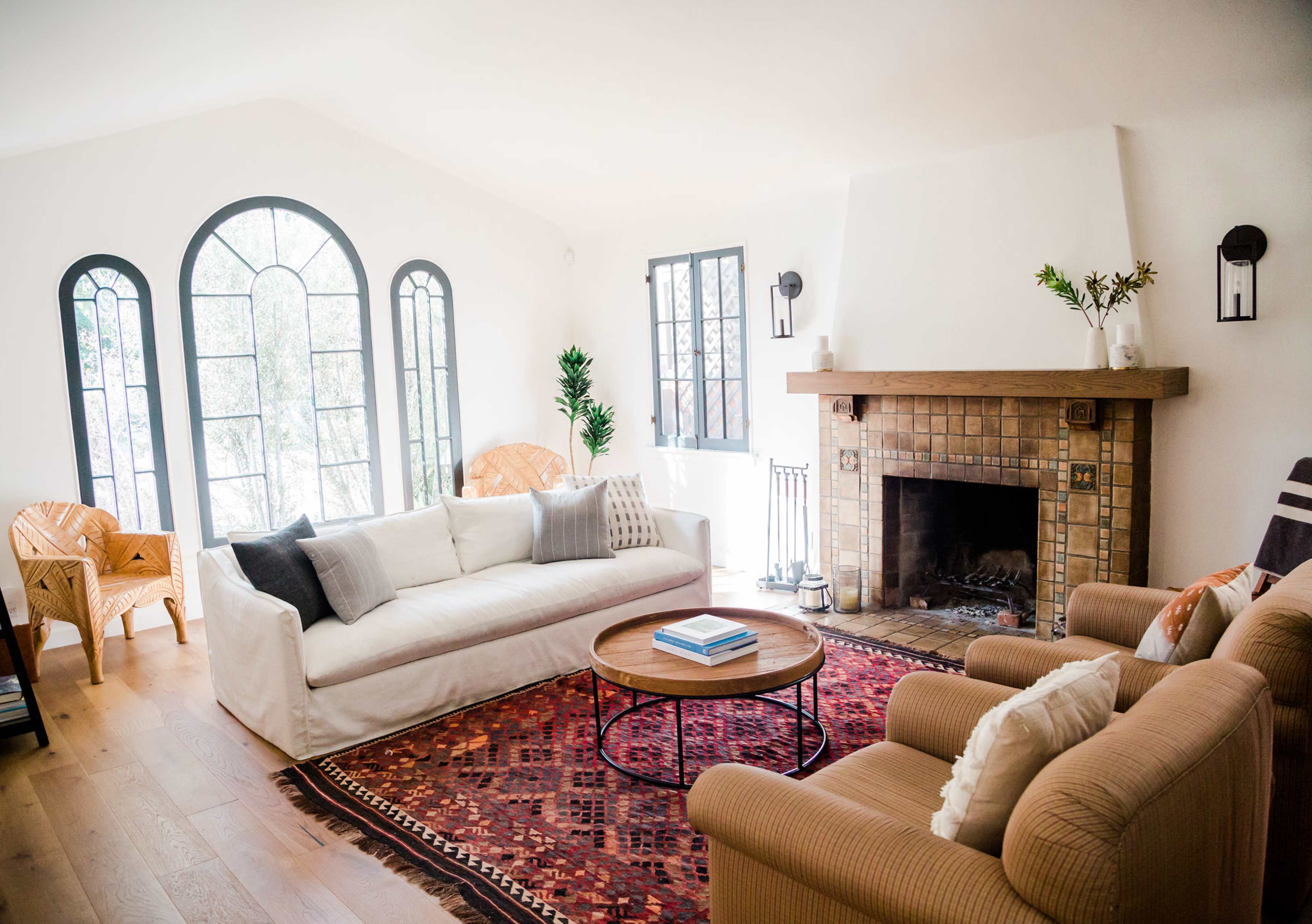A bright living room features a white sofa, two tan armchairs, a round coffee table, and a tiled fireplace, all set against large arched windows and a colorful area rug.
