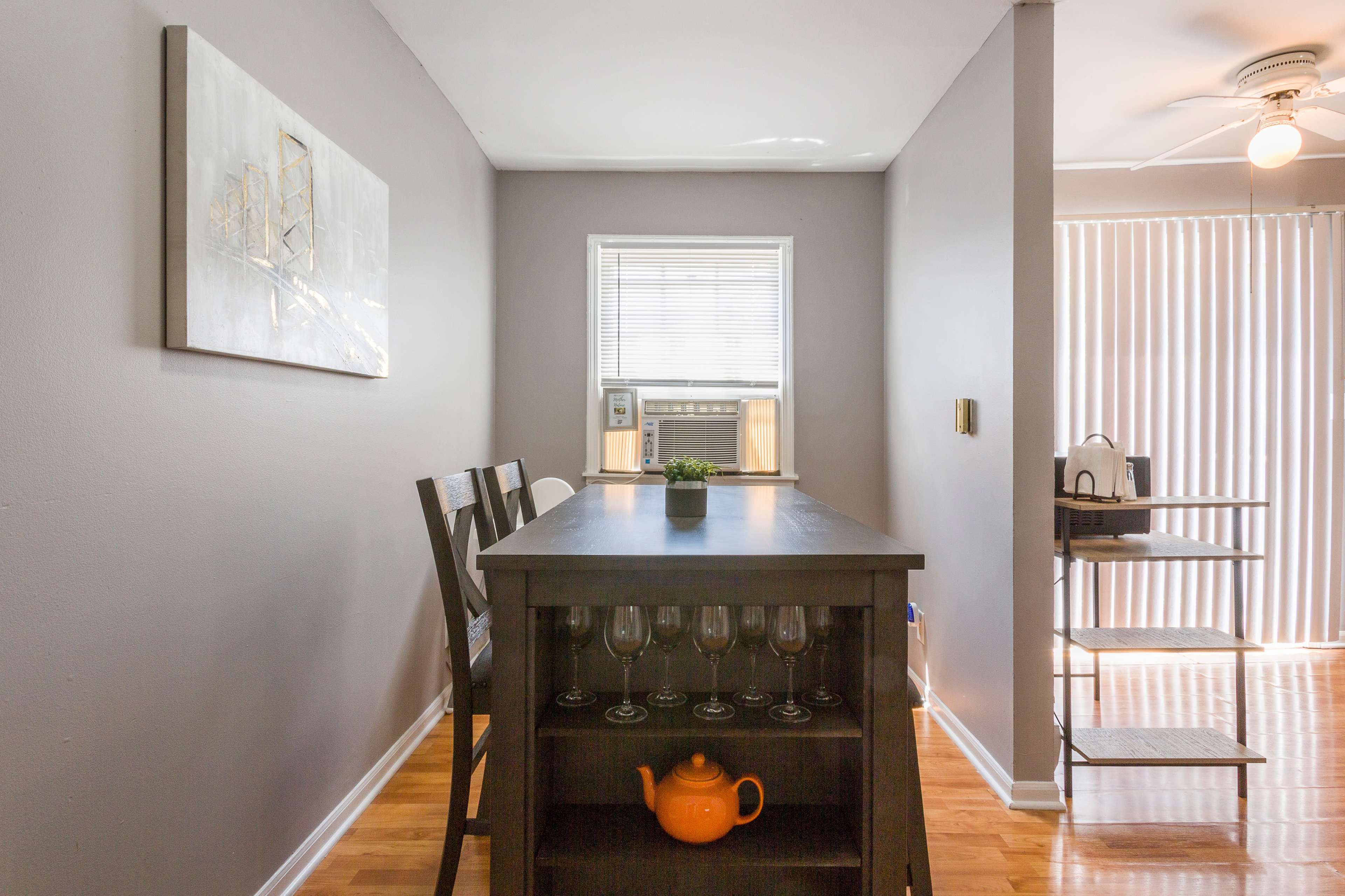 A simple dining area with a wooden table, four chairs, and a small shelf with a teapot and glasses, set against gray walls and a window with blinds.