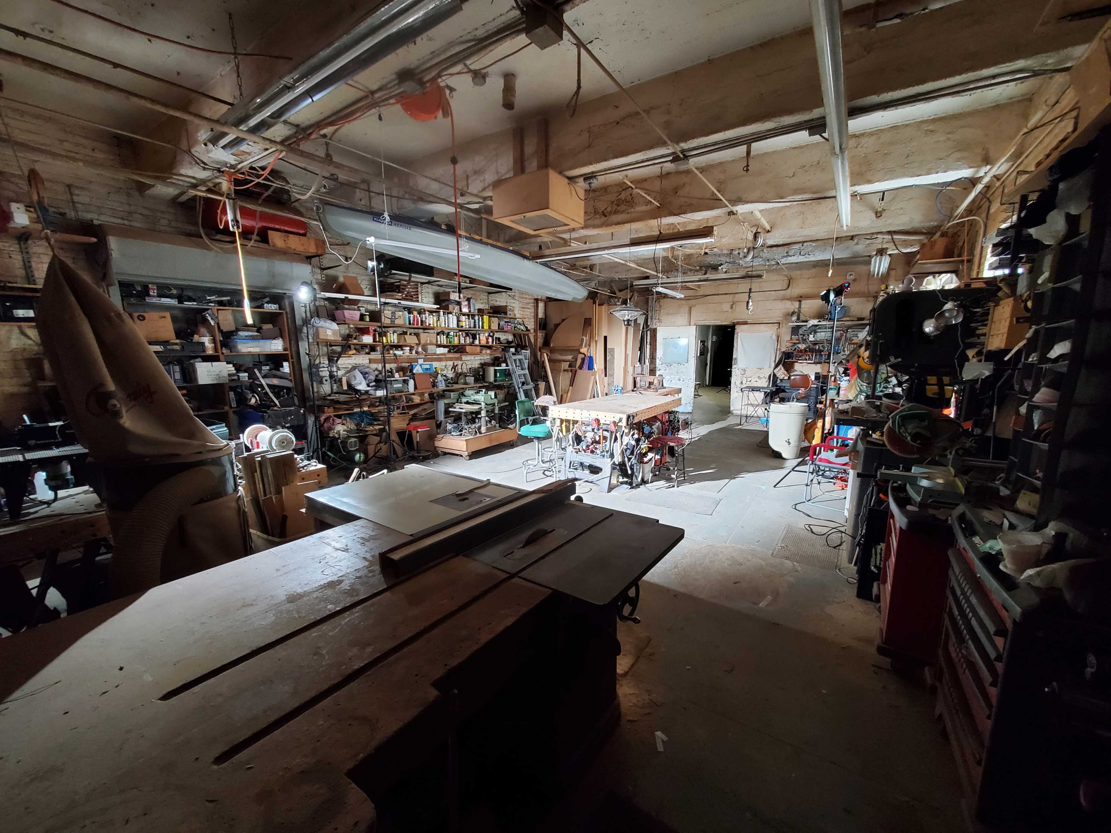 A cluttered workshop filled with tools, equipment, and workstations, illuminated by overhead lighting.