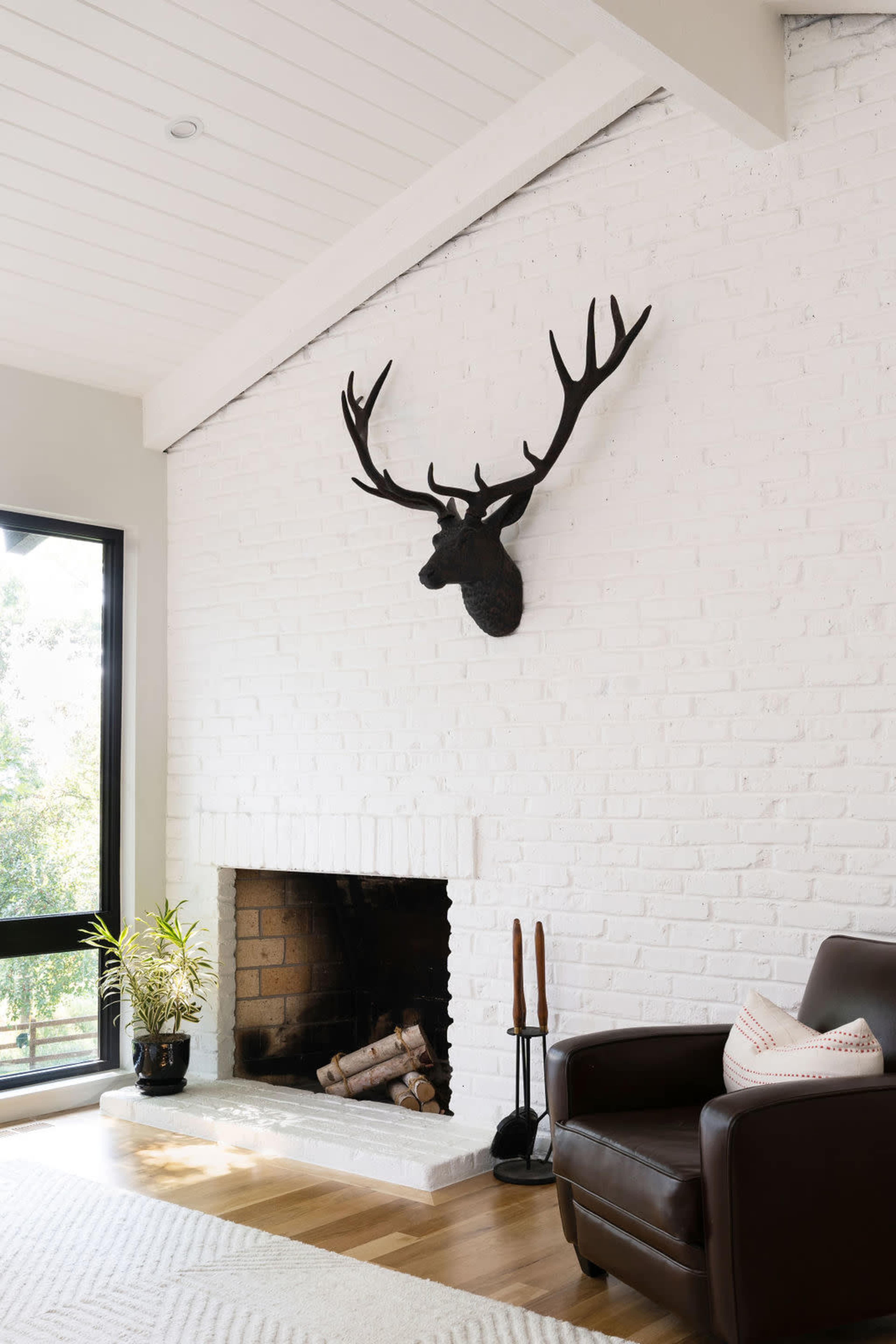 The image shows a modern living room featuring a white brick fireplace with a mounted deer head above it, a leather armchair to the side, and a small plant in a pot.