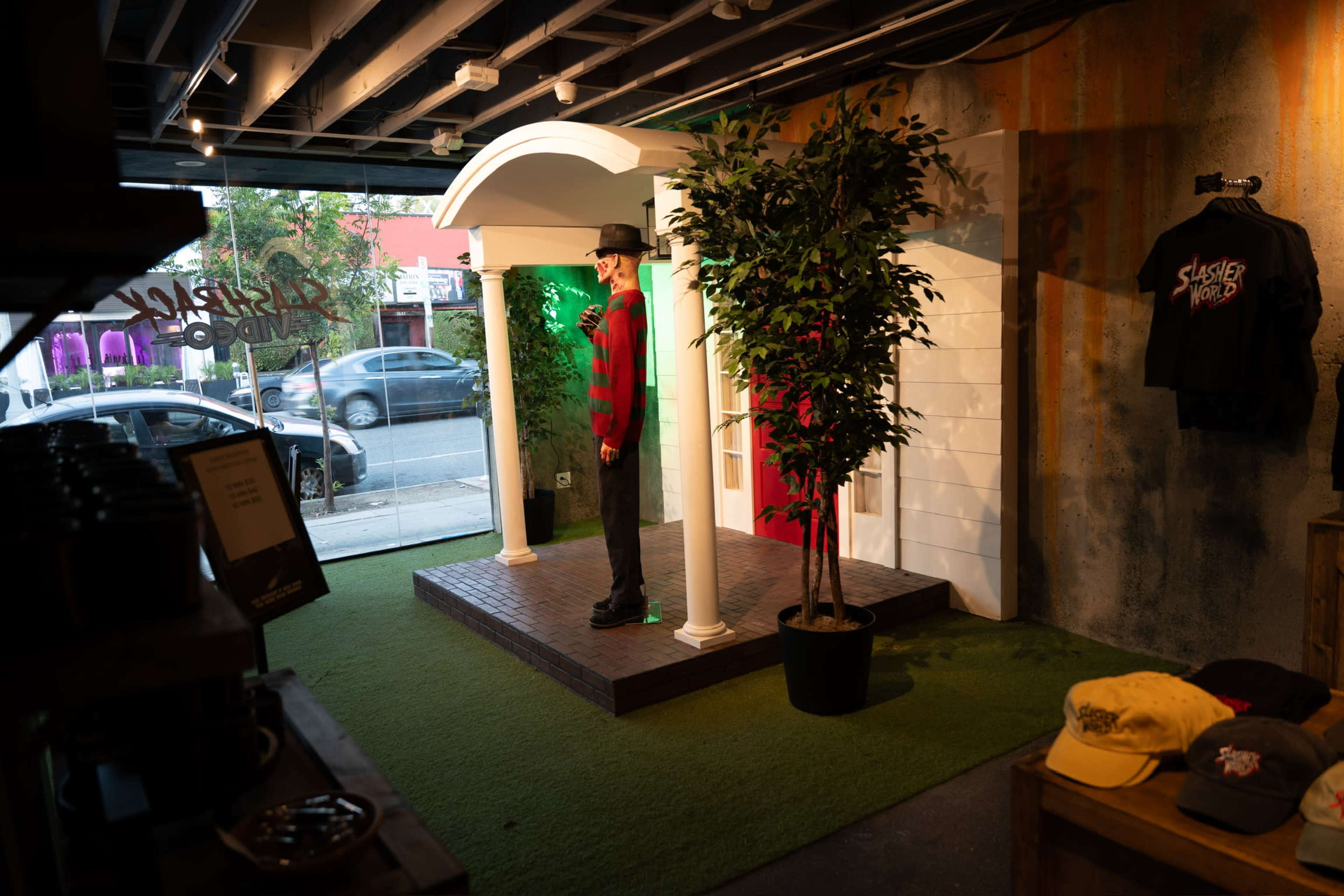 The image shows a display setup with a mannequin dressed in a striped outfit under a white columned canopy, surrounded by artificial greenery and merchandise on a shelf.