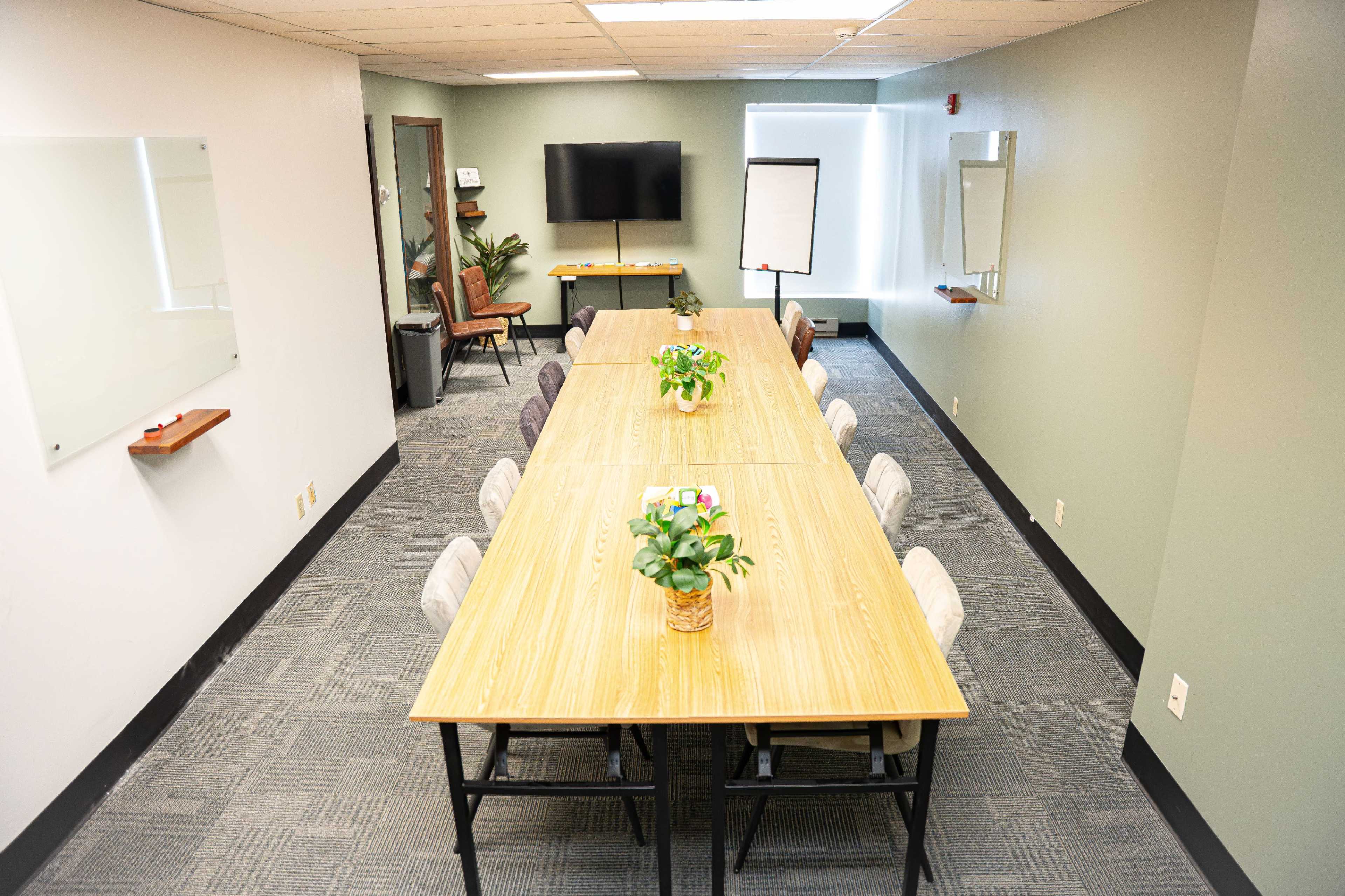 The image shows a conference room with a long wooden table, surrounded by chairs, and equipped with a television, whiteboard, and potted plants.