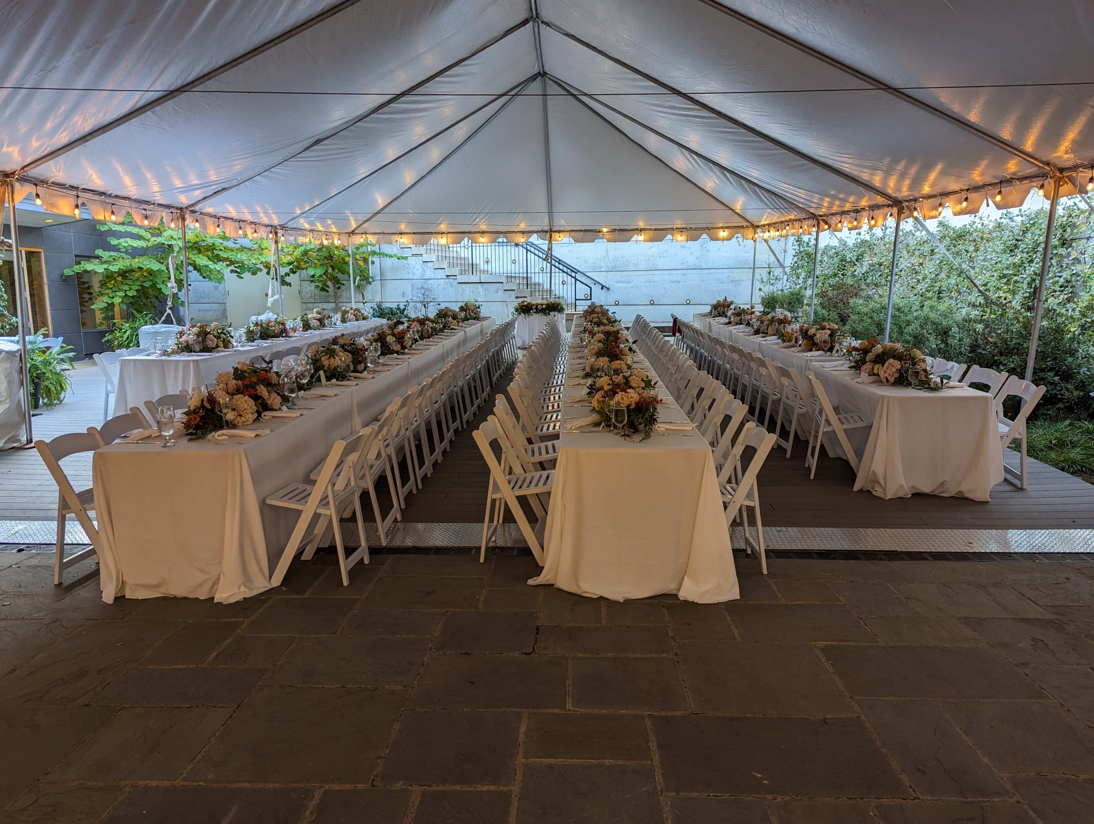 A large outdoor tent is set with multiple tables covered in white tablecloths, arranged in two parallel lines, each adorned with flower centerpieces.