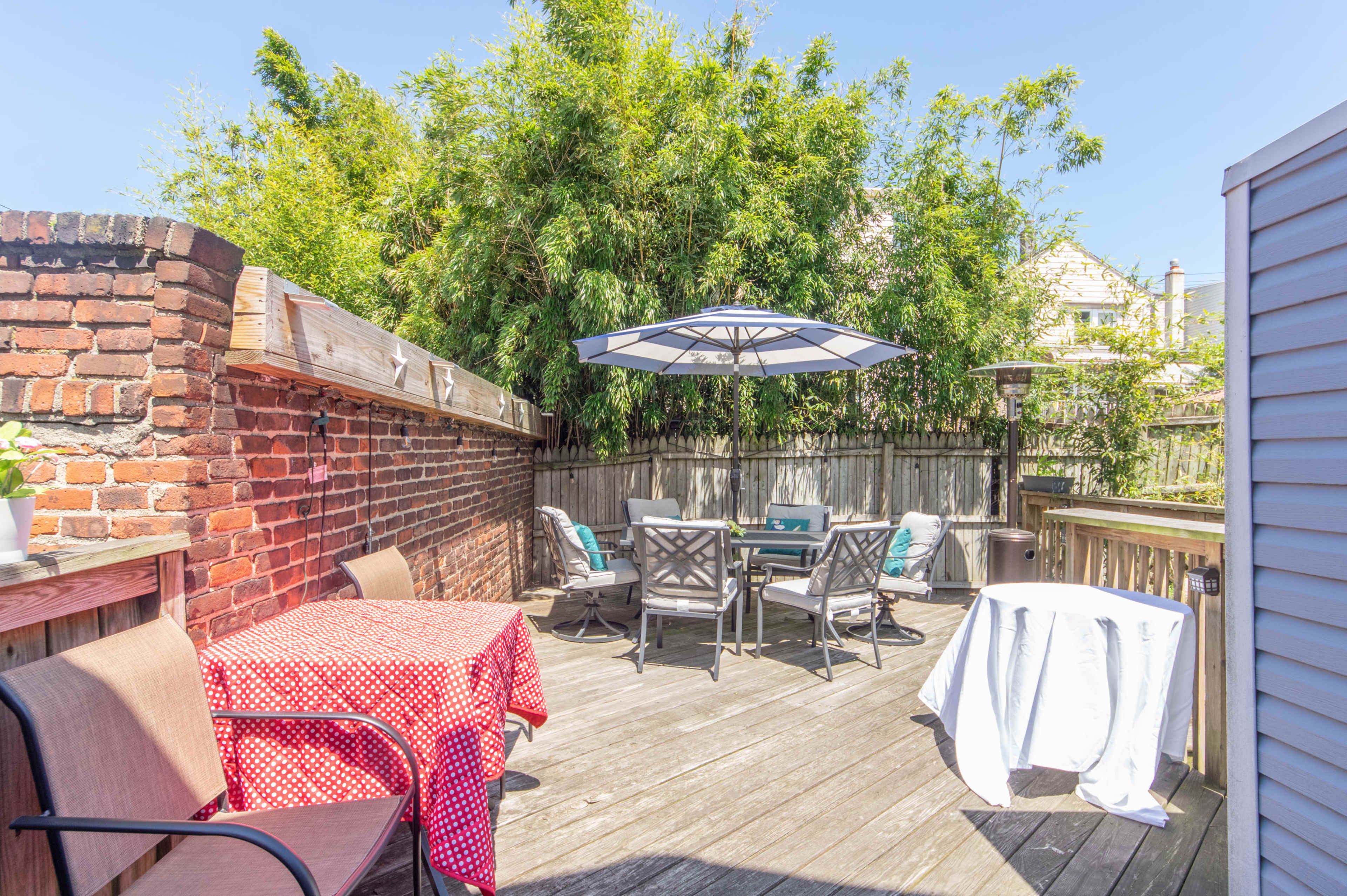A wooden deck features a red-and-white checkered tablecloth on one table, outdoor seating, an umbrella, and a backdrop of green bamboo plants.