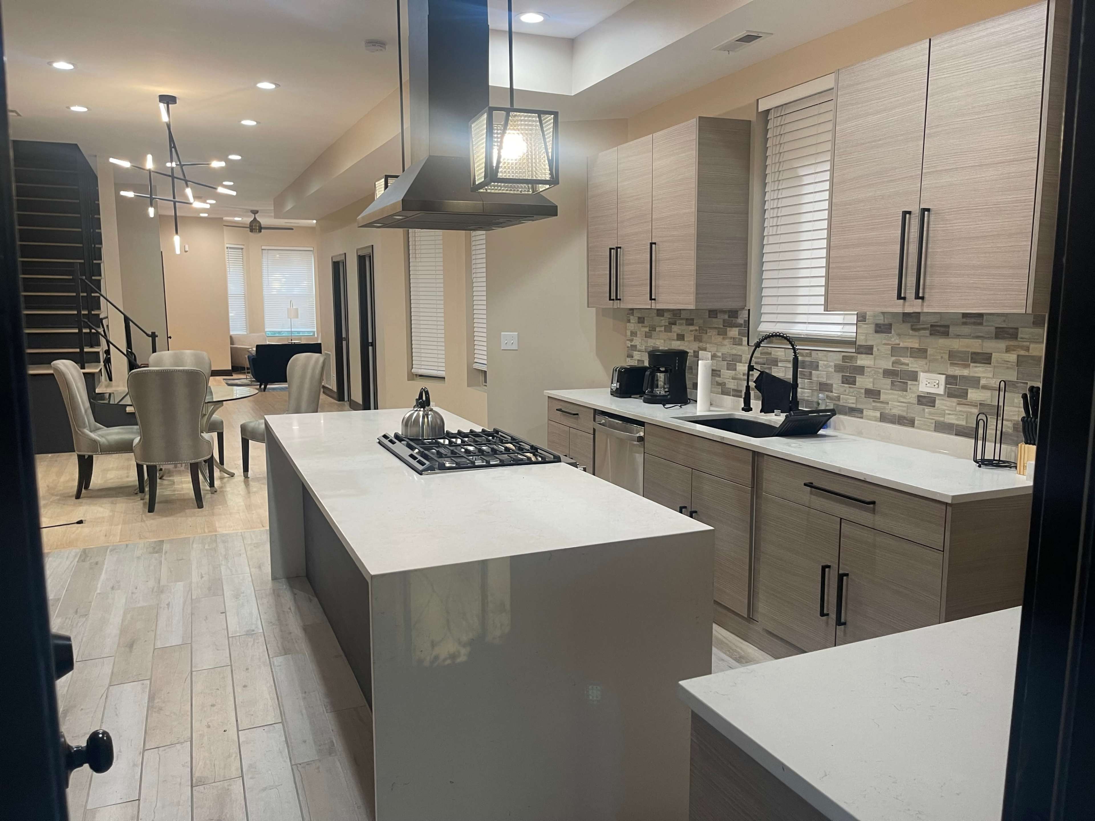 A modern kitchen with light wood cabinetry, a large island with a gas cooktop, and a view into a dining area with chairs in the background.
