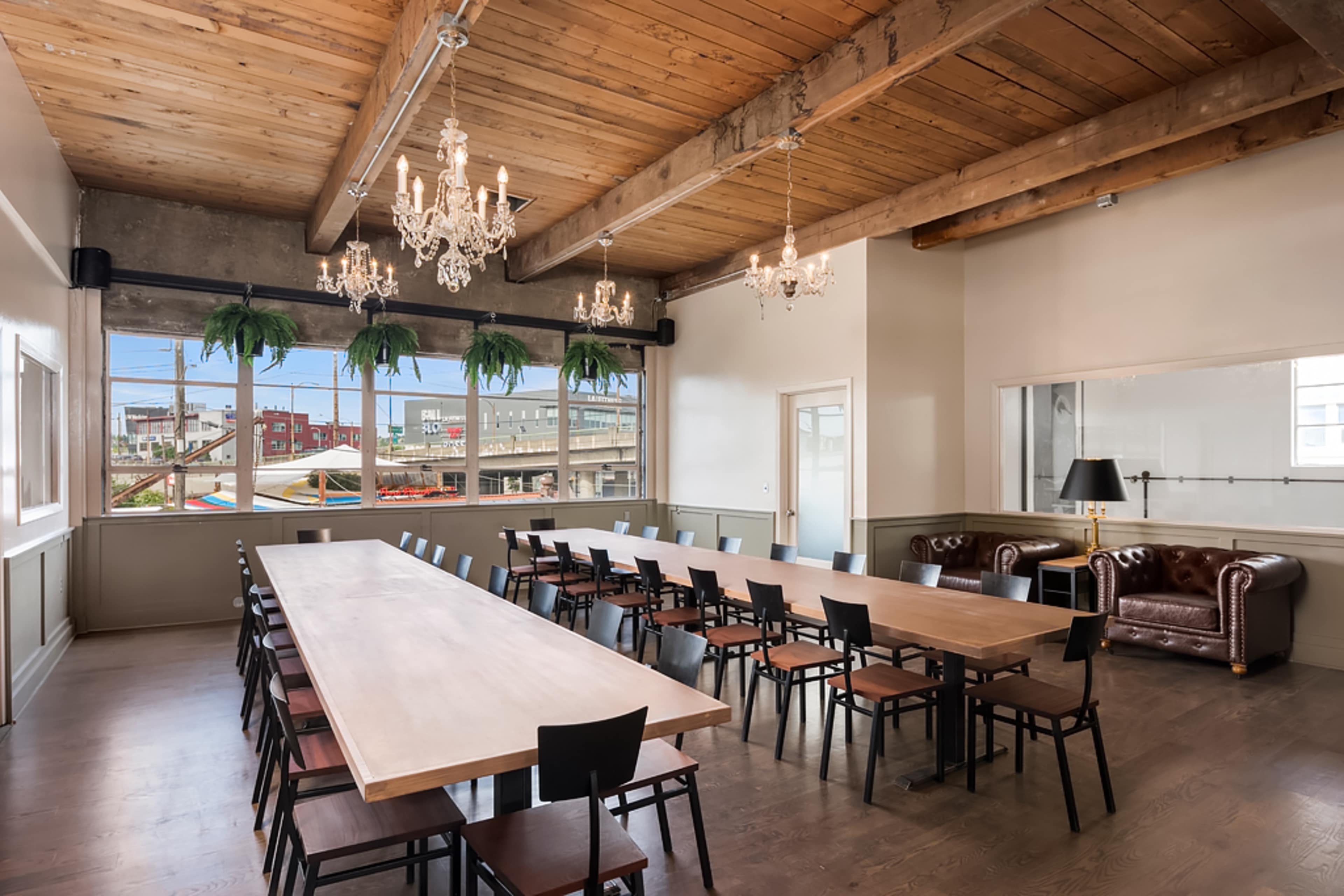 A large meeting room features a long wooden table surrounded by chairs, with chandeliers hanging from the ceiling and large windows providing natural light.