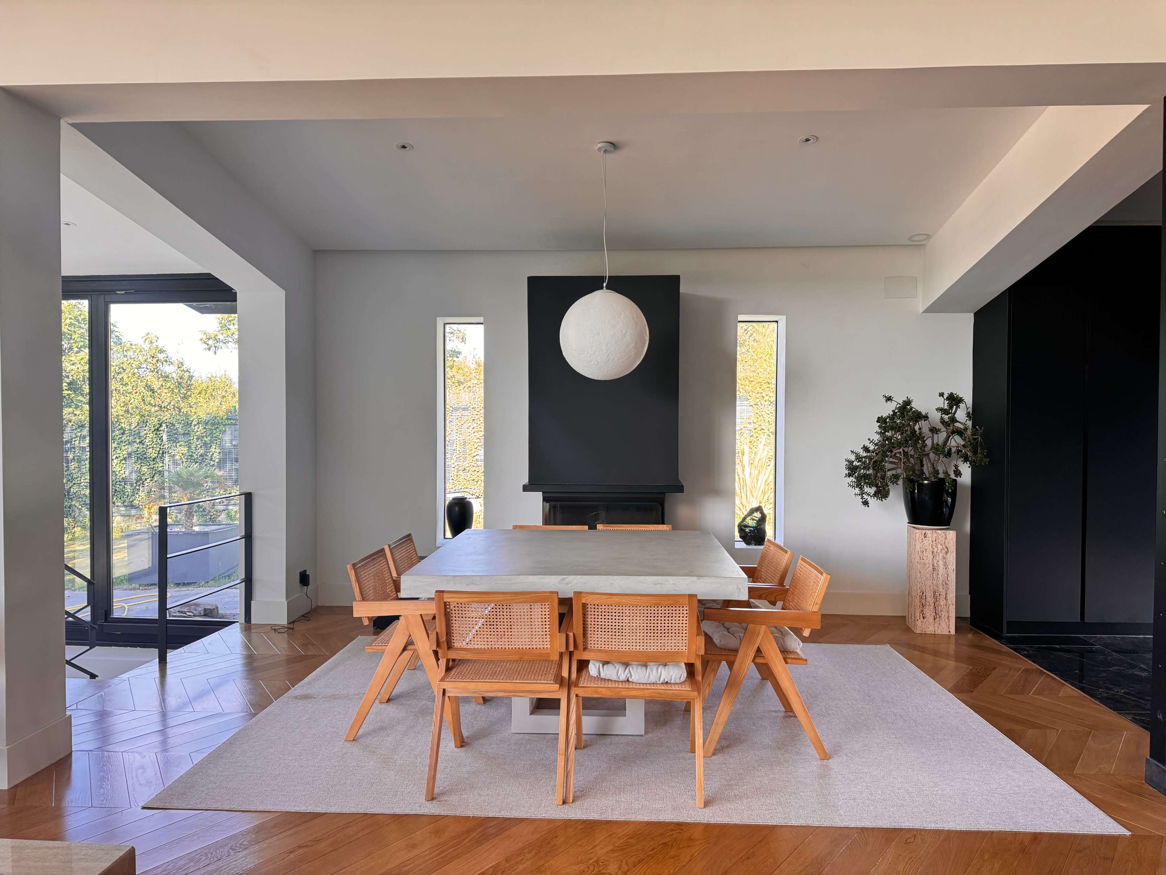 The image shows a modern dining room with a large rectangular table surrounded by wooden chairs, a pendant light above, and large windows allowing natural light to enter.