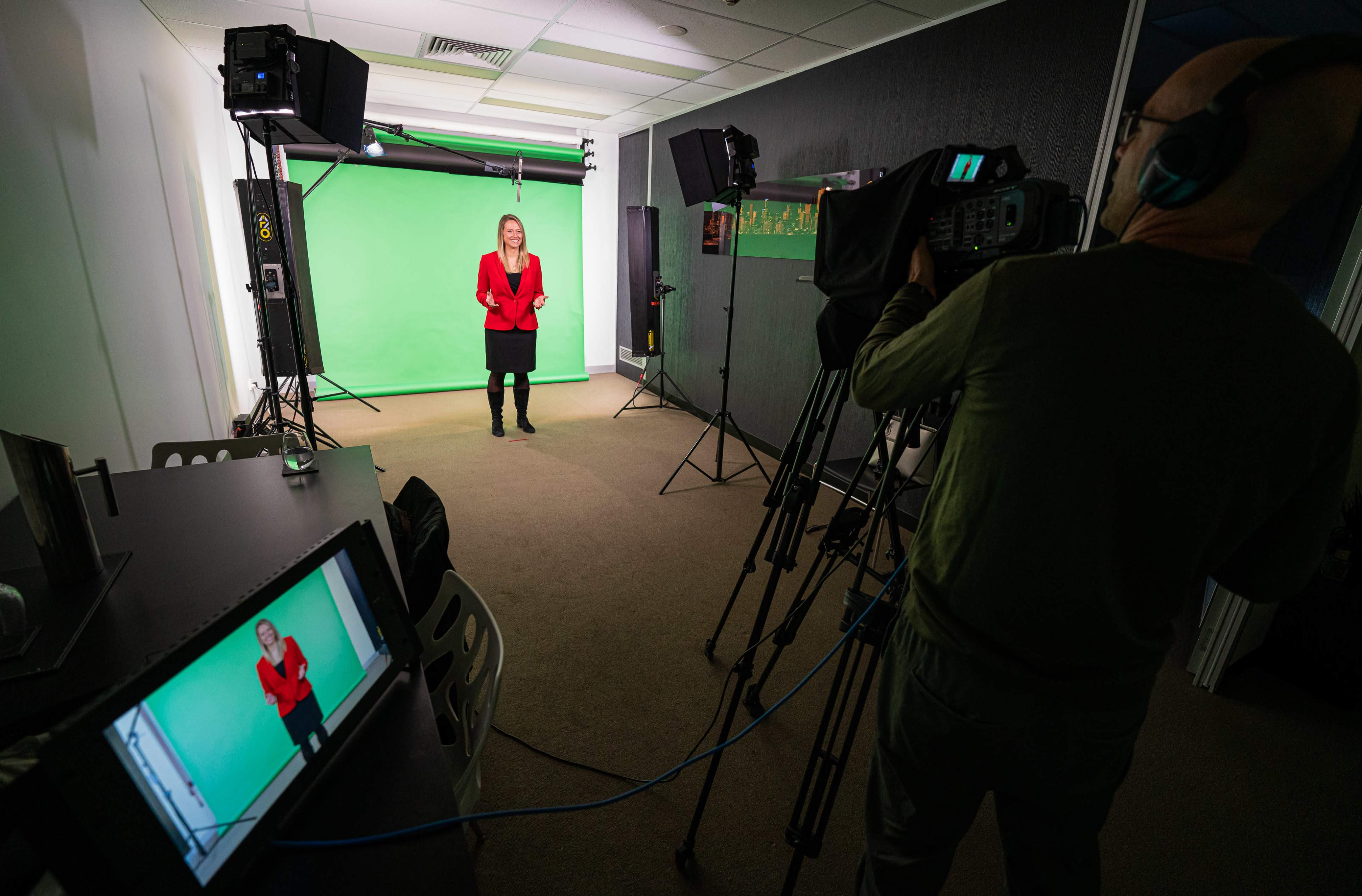 A woman in a red blazer stands in front of a green screen, with a camera and crew capturing her from the opposite side of the room.