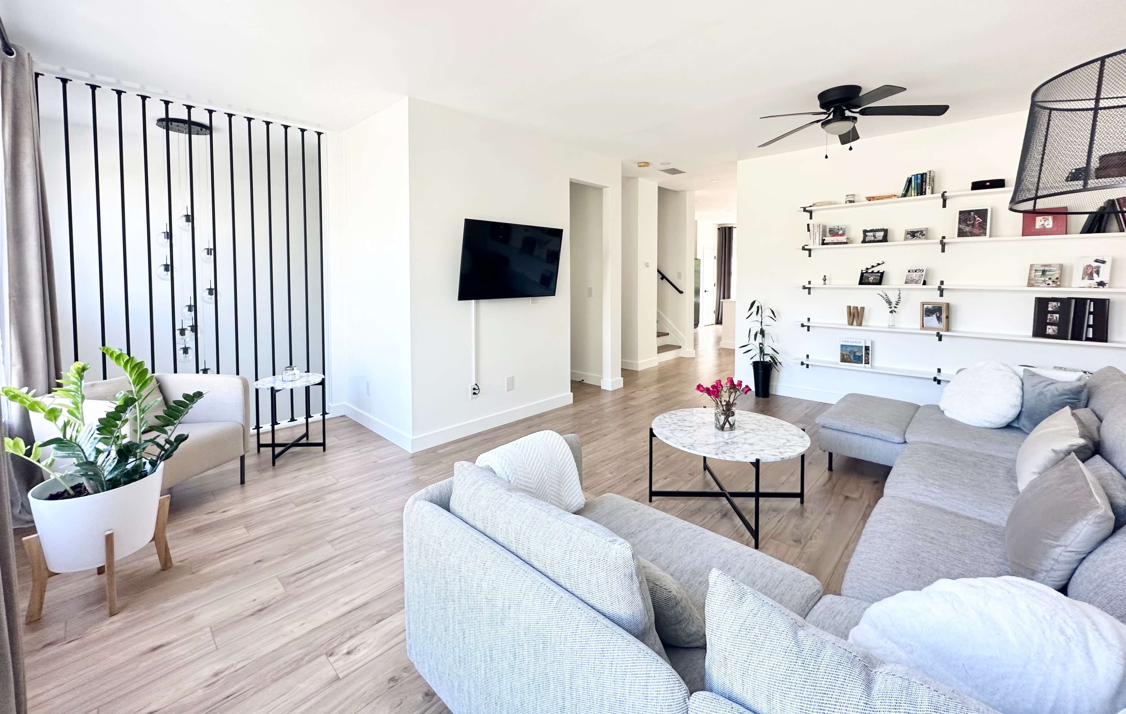 The living room features a light-colored sectional sofa, a round coffee table, a television mounted on the wall, and shelves displaying various items.