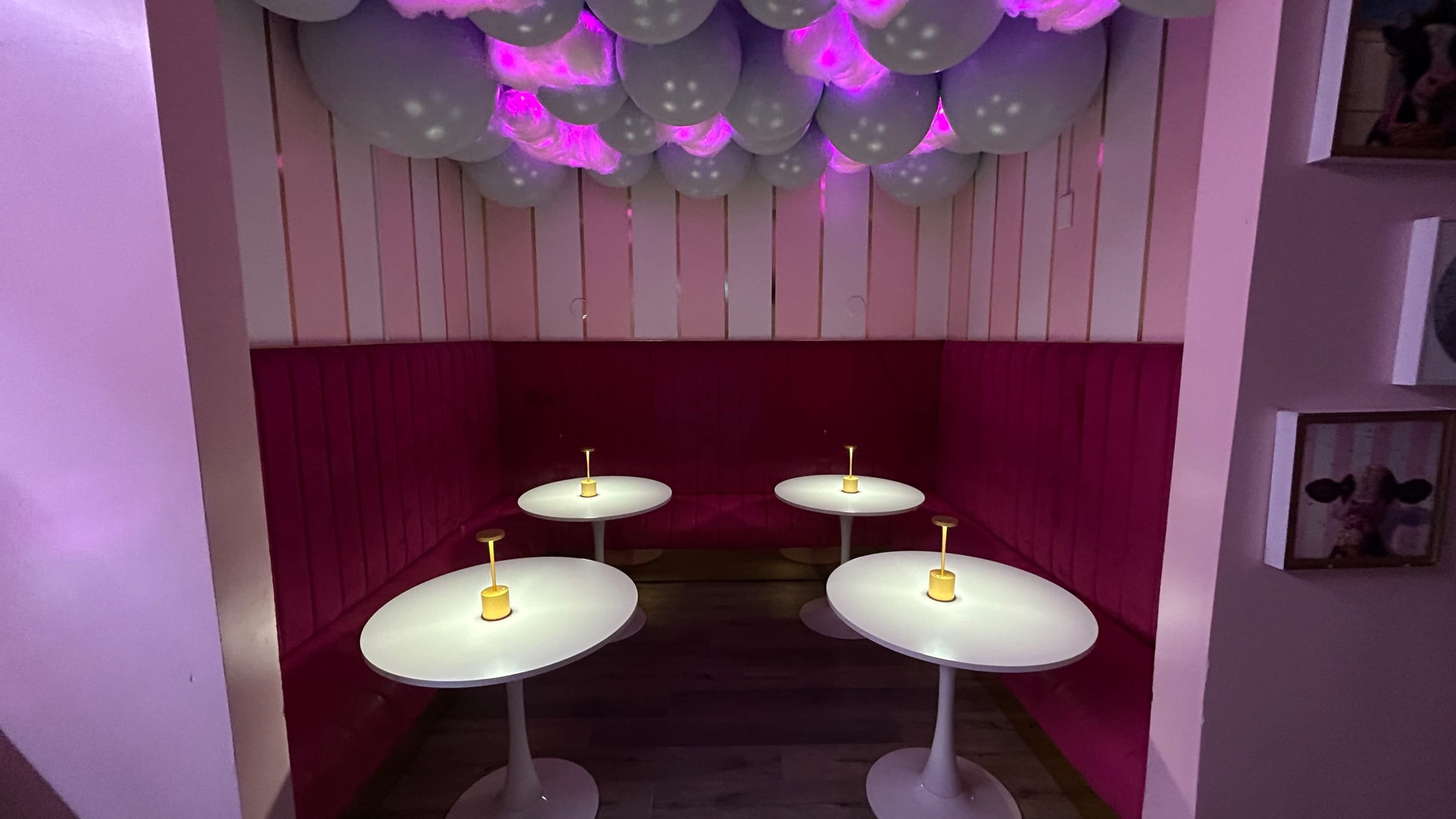 A cozy booth area with four white tables topped with candles, surrounded by pink walls and a ceiling adorned with balloons lit in purple hues.