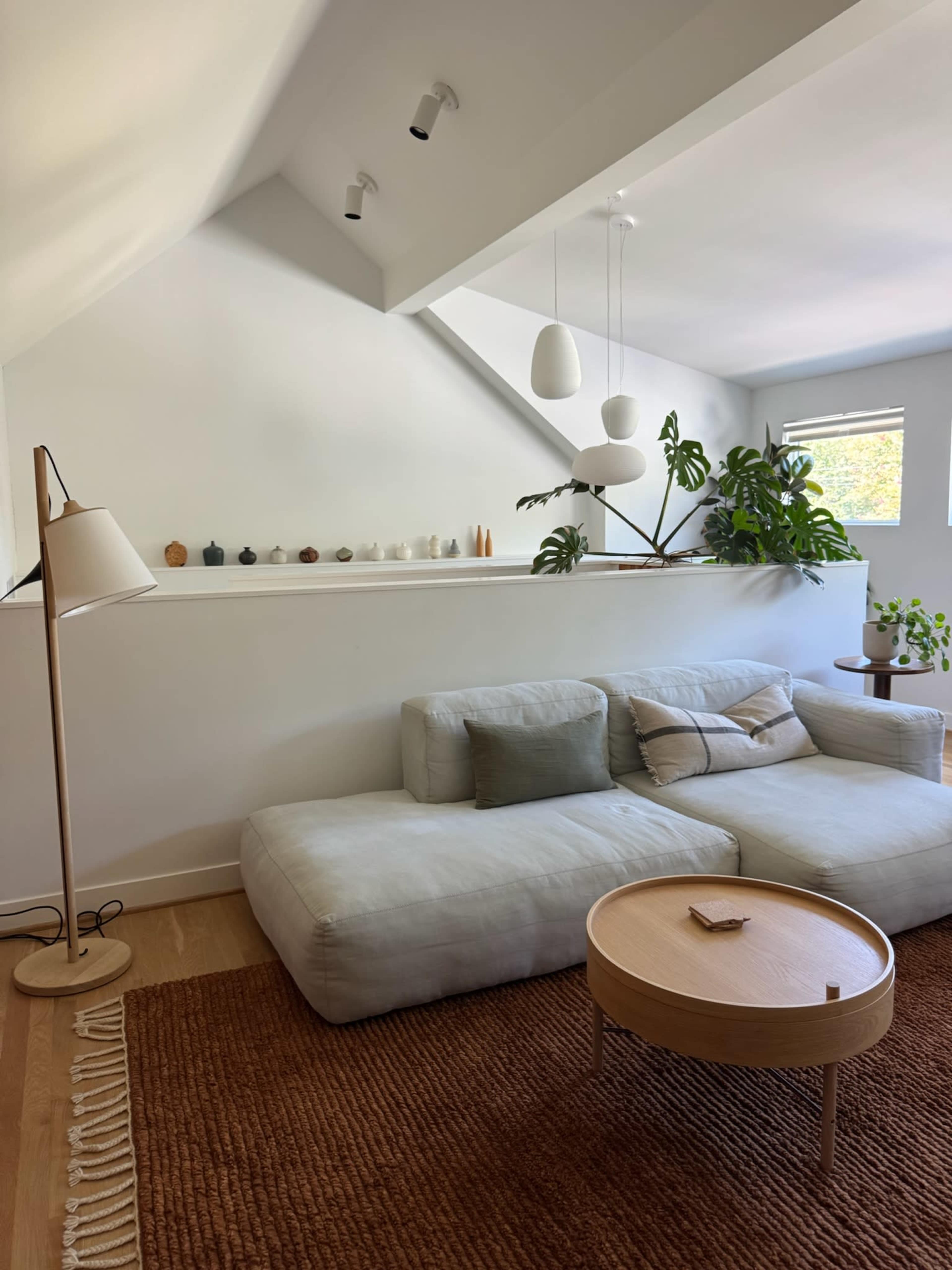 The image shows a modern living room with a large, light-colored sofa, a round wooden coffee table, and plants placed around the space.