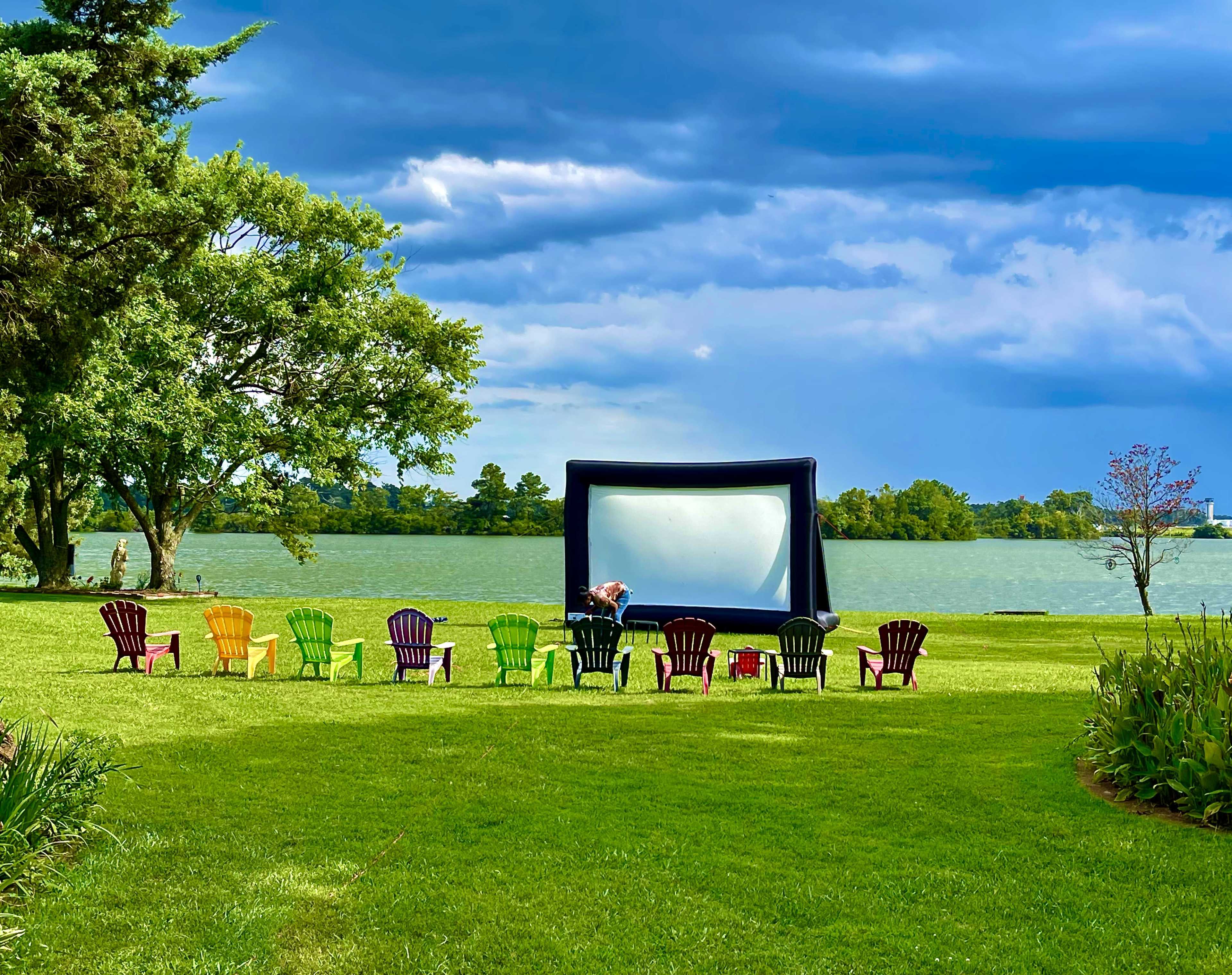 A large inflatable movie screen is set up near a lake, surrounded by colorful Adirondack chairs on a grassy area.