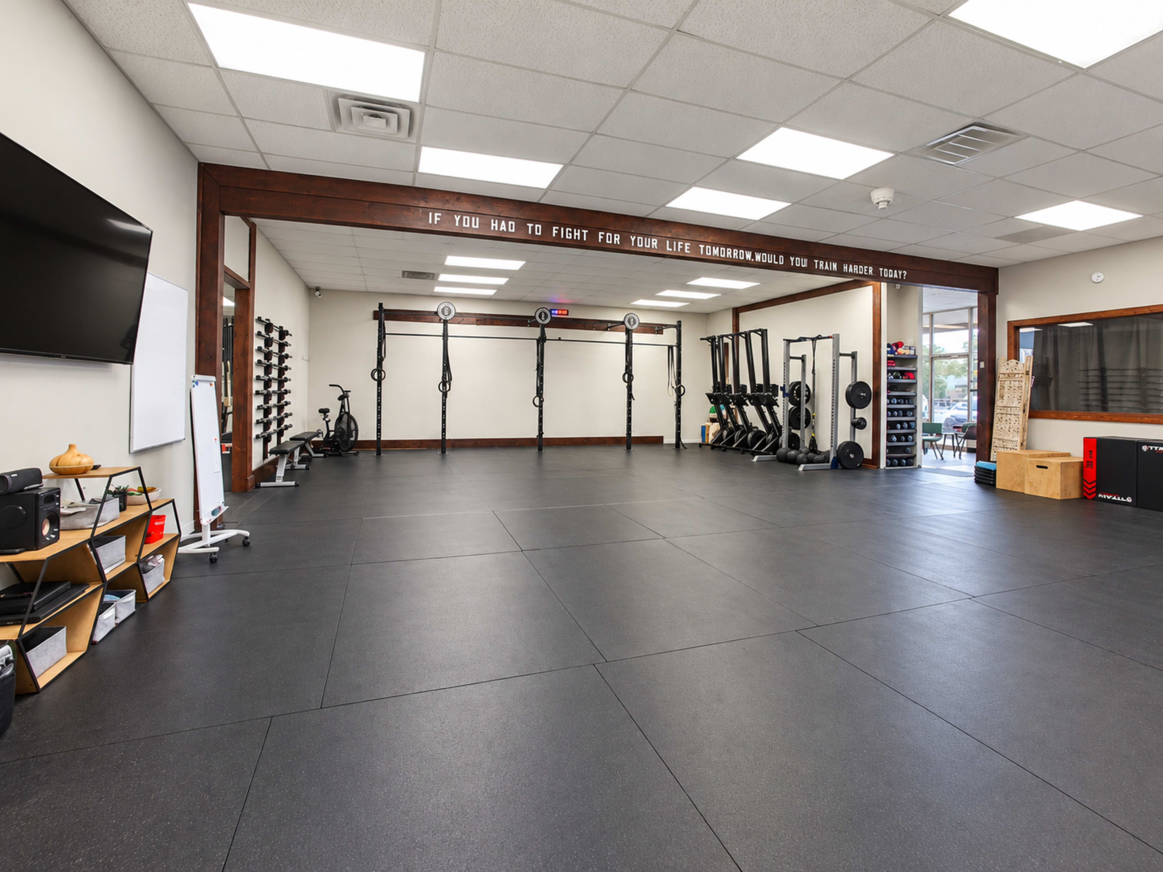 The image shows a spacious gym interior with exercise equipment, a large open floor, and motivational text on the wall.