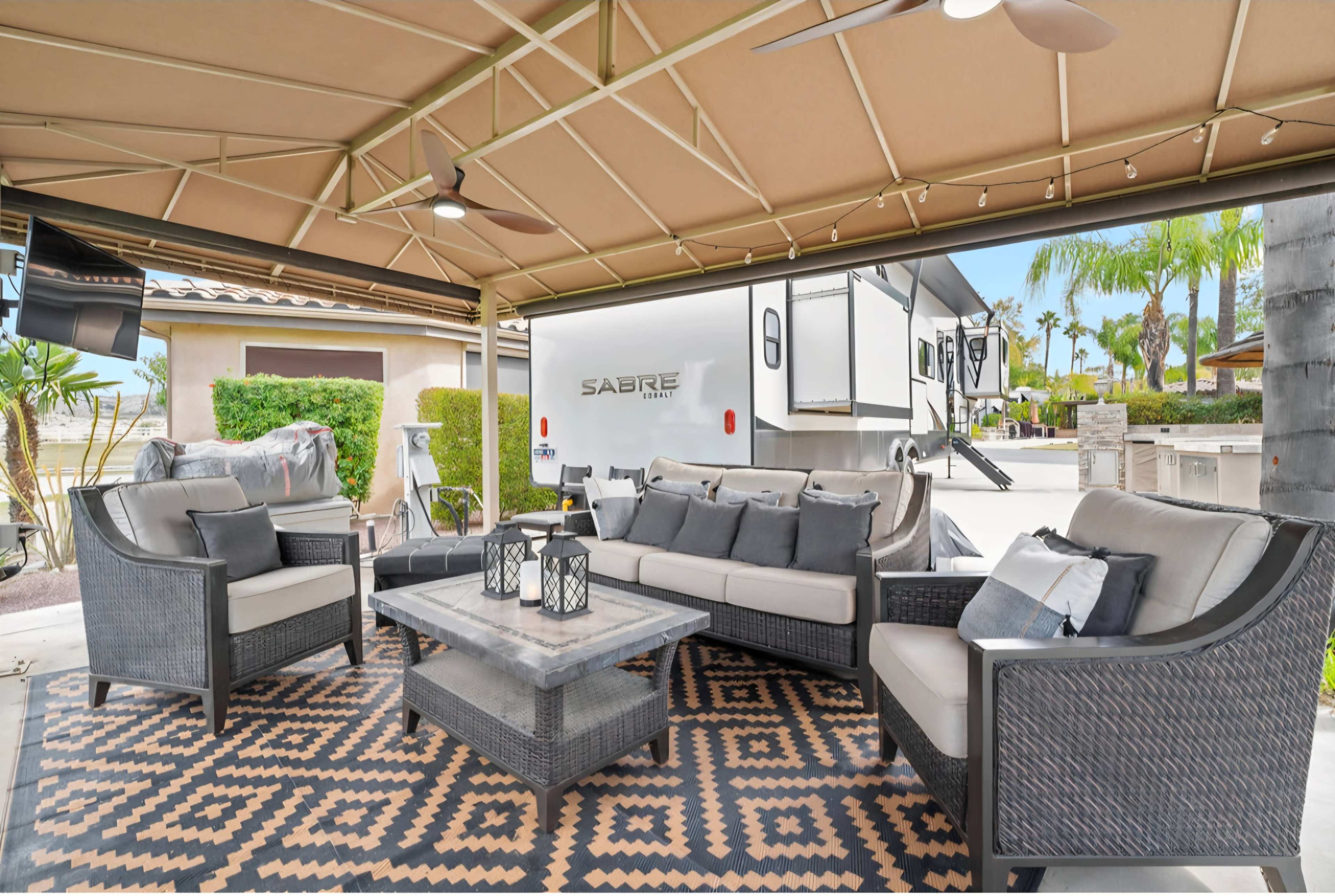 The image shows a covered outdoor seating area with a sectional sofa, coffee table, and decorative lanterns, set against a backdrop of a trailer and palm trees.