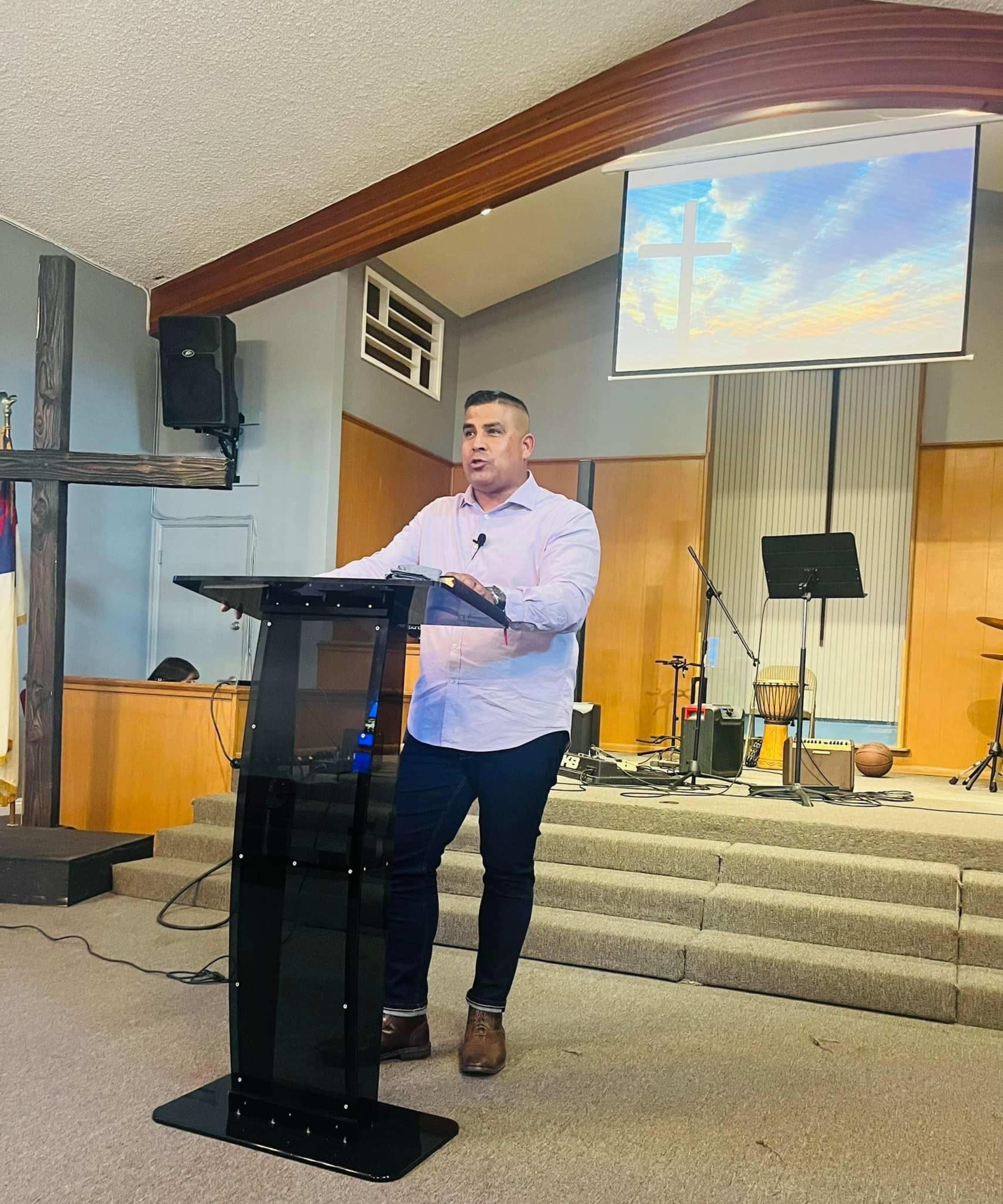A man stands behind a clear pulpit in a church, speaking to the congregation with a projector screen displaying a cross in the background.