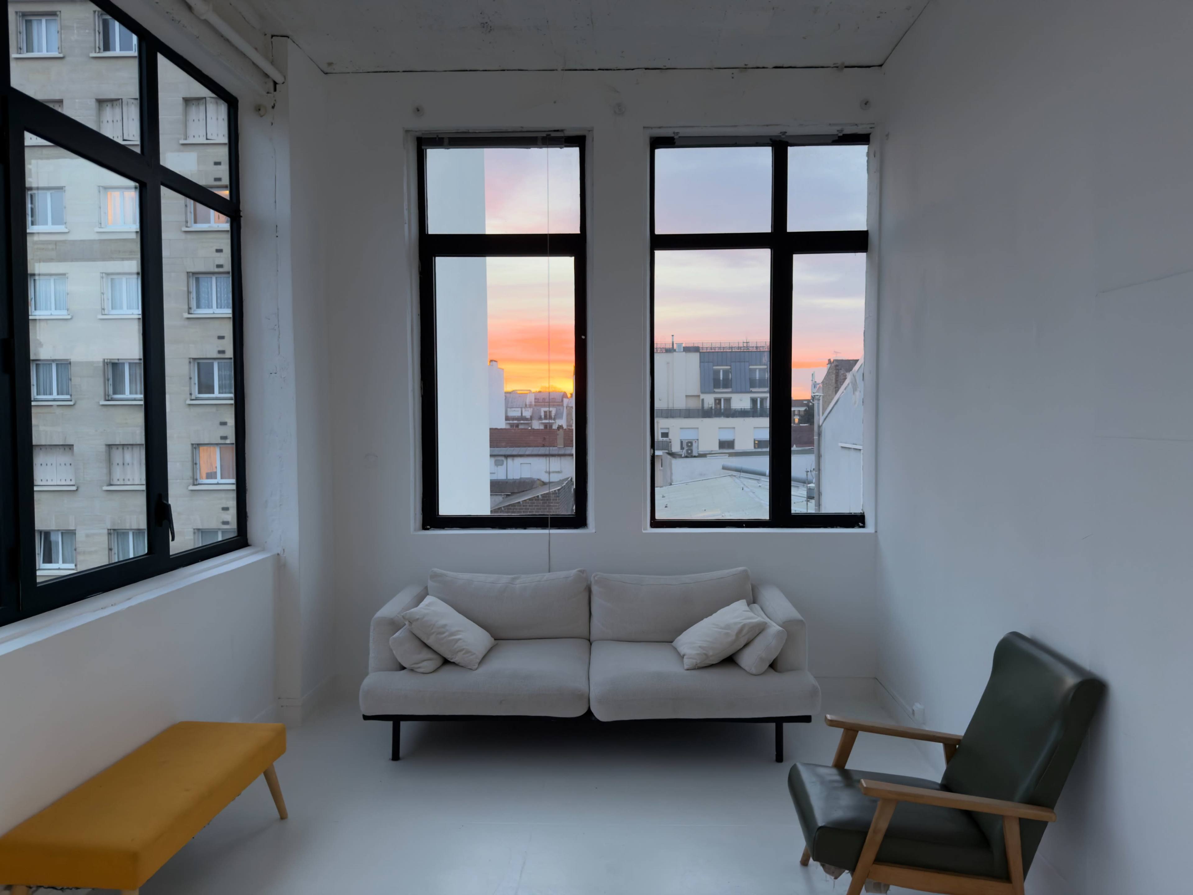 A minimalist living room features a light-colored sofa and a green armchair, with large windows showcasing a vibrant sunset over a cityscape.