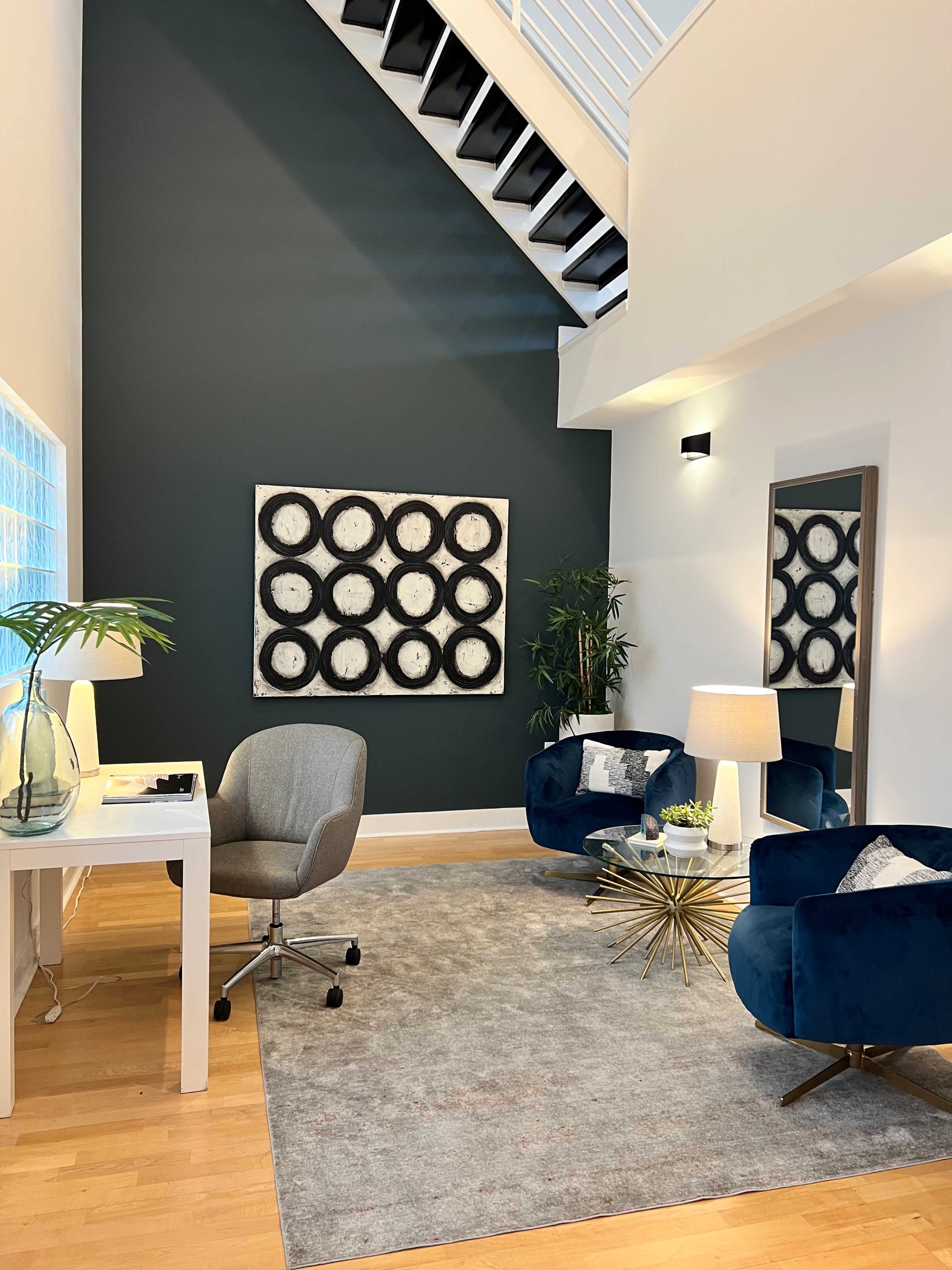 A modern interior space with a dark green accent wall, featuring a desk, a swivel chair, two blue armchairs, a coffee table with gold legs, and a large mirror.