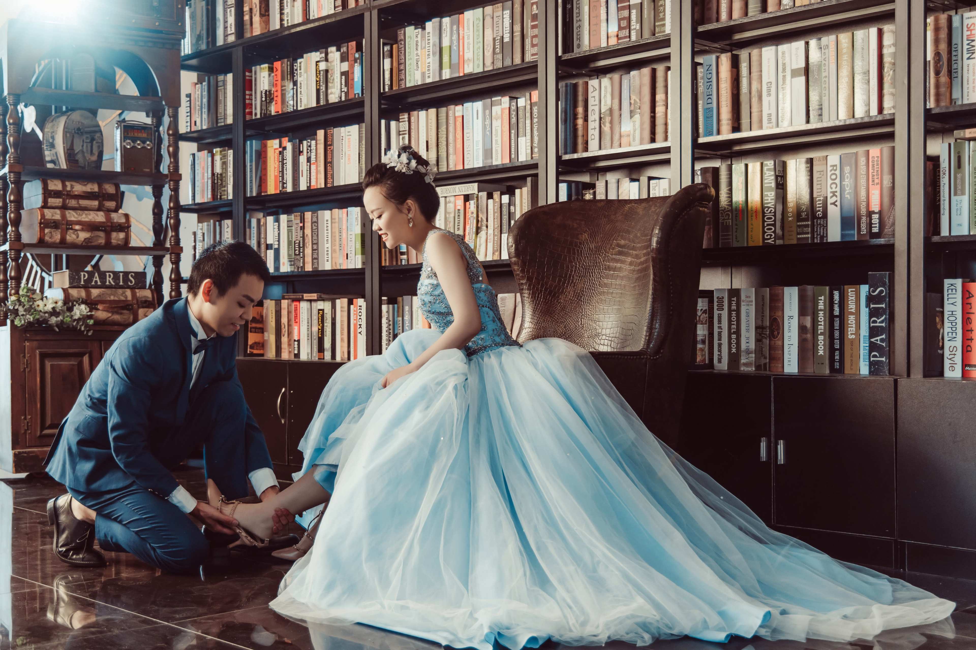 A man kneels to help a woman wear a shoe while seated in front of a large bookshelf filled with books.